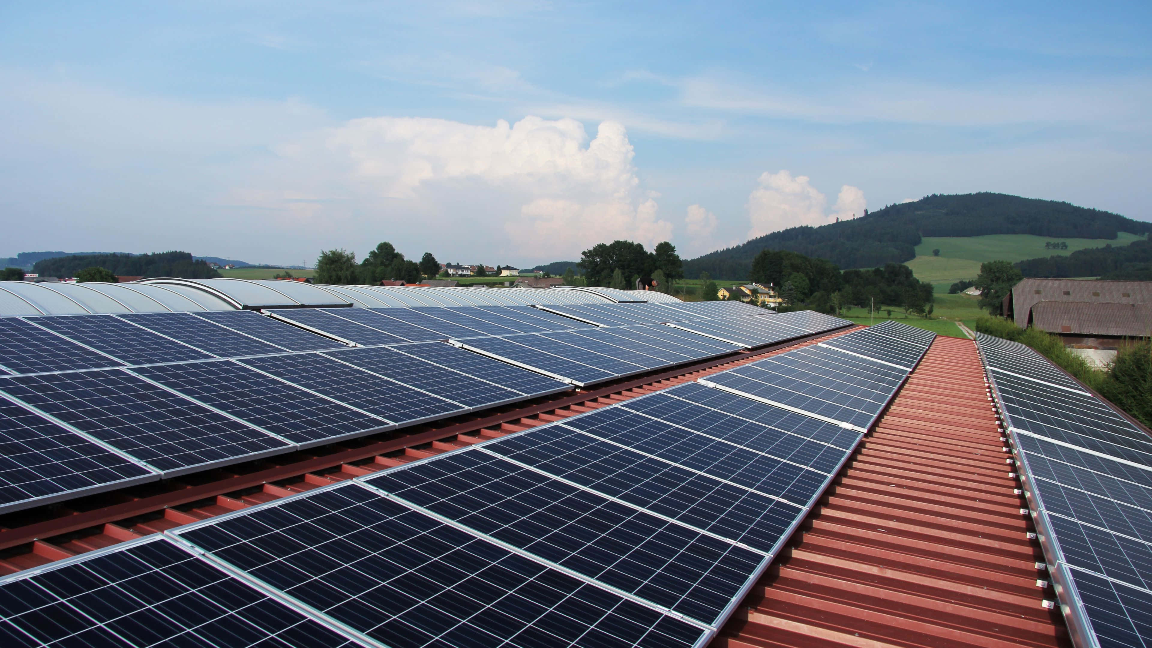 Solar Panel 桌布 壁紙 — 一排排太陽能板在明亮的藍天和蓬鬆白雲下延伸在紅色屋頂上。