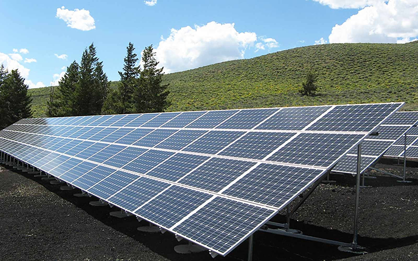 Solar Panel 桌布 壁紙 — 一排排太陽能板在晴朗藍天白雲下閃閃發光，背景是綠色山坡和松樹。