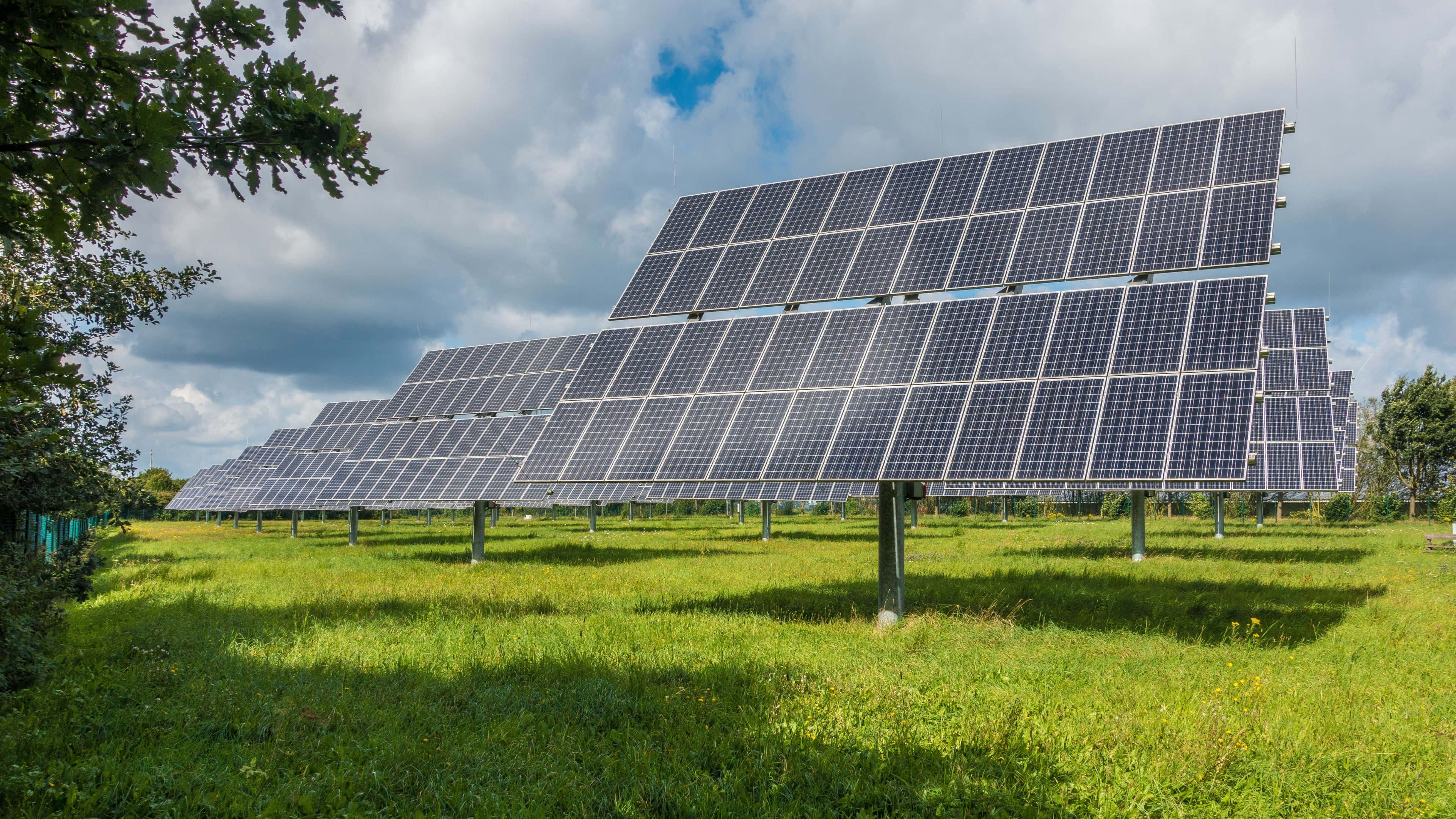 Solar Panel 桌布 壁紙 — 一排排太陽能板在陽光明媚的綠色田野上延伸，天空多雲，藍天白雲。