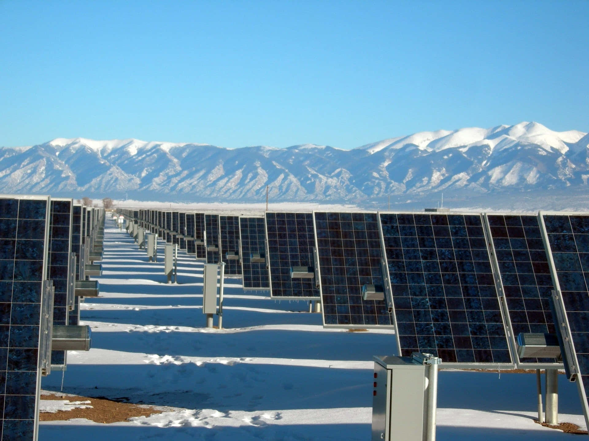 Electricity 桌布 壁紙 — 一排排太陽能板延伸在雪地上，背景是白雪覆蓋的山脈，天空清澈湛藍。