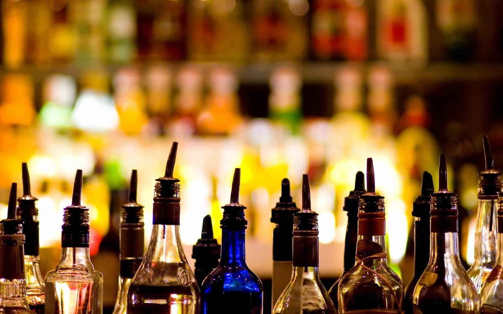 Drink 桌布 壁紙 — 一排排帶有倒酒嘴的酒瓶在溫暖模糊的酒吧層架上清晰對焦，散發著琥珀色的光芒。