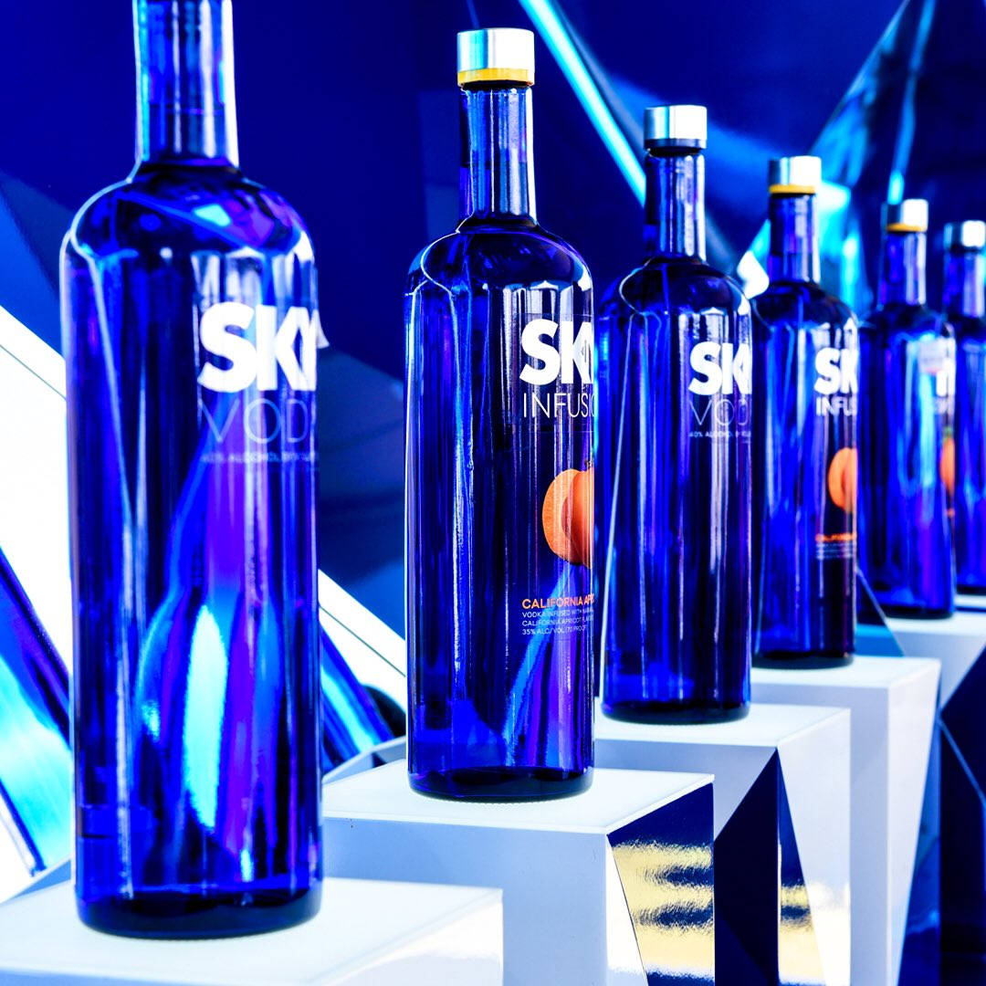 alcohol 桌布 壁紙 — 一排排帶有橙色標籤的光澤藍色SKI伏特加酒瓶，在充滿活力的藍色燈光下矗立在白色幾何底座上。