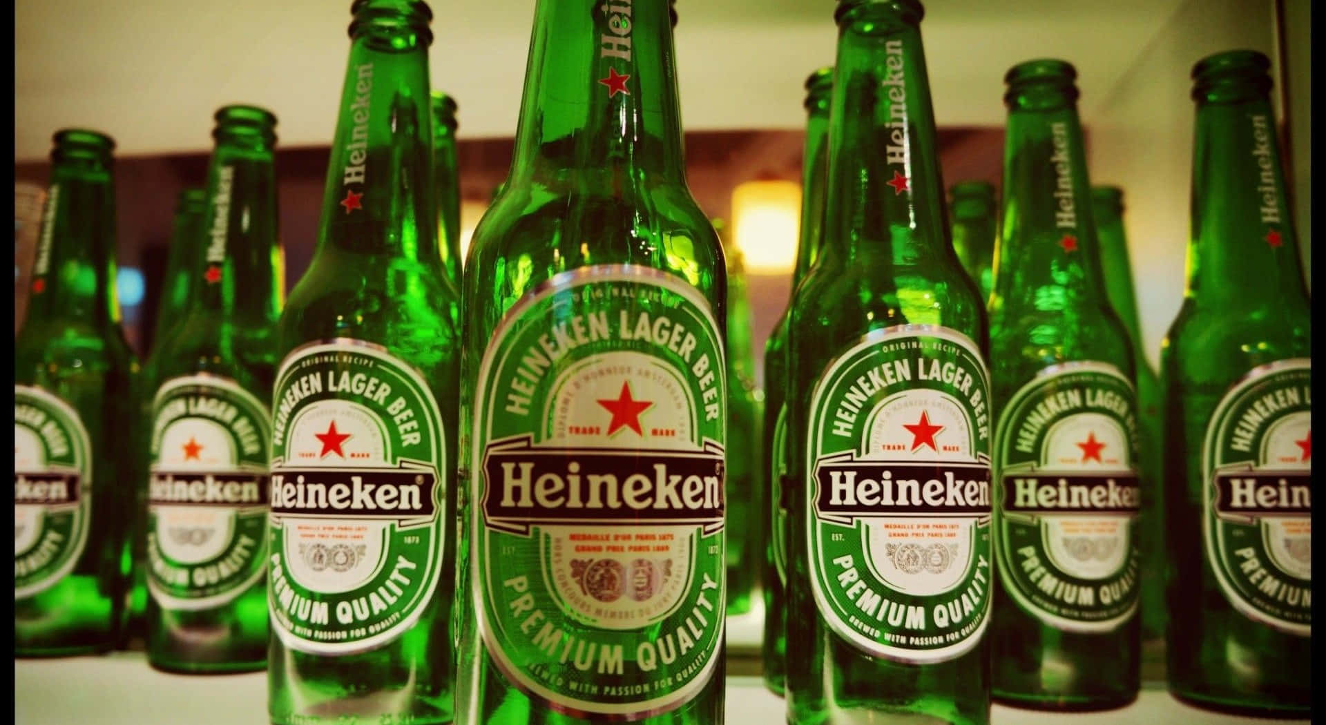 Heineken 桌布 壁紙 — 一排排帶有紅色星星標籤的綠色海尼根啤酒瓶，在溫暖的室內燈光下陳列。