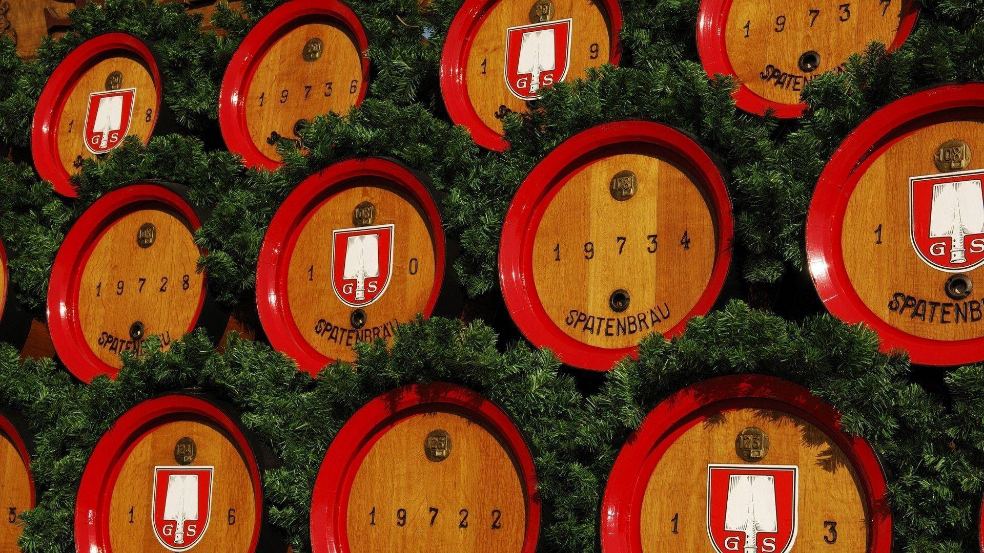 慕尼黑啤酒節 桌布 壁紙 — 一排排帶有紅色邊緣和 'Spatenbrau' 標誌的木製啤酒桶，藏在綠色松樹枝之間。