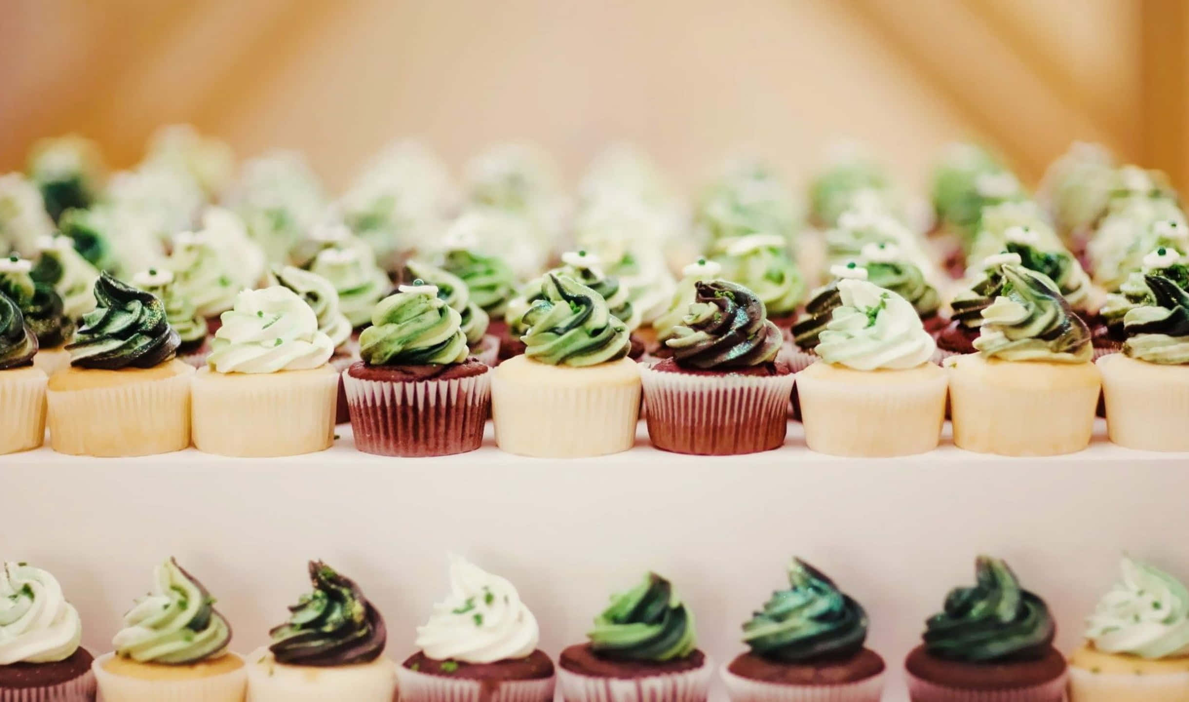 2440x1440 Desserts 桌布 壁紙 — 一排排帶有綠色和白色糖霜漩渦的杯子蛋糕整齊地放在白色多層架上。
