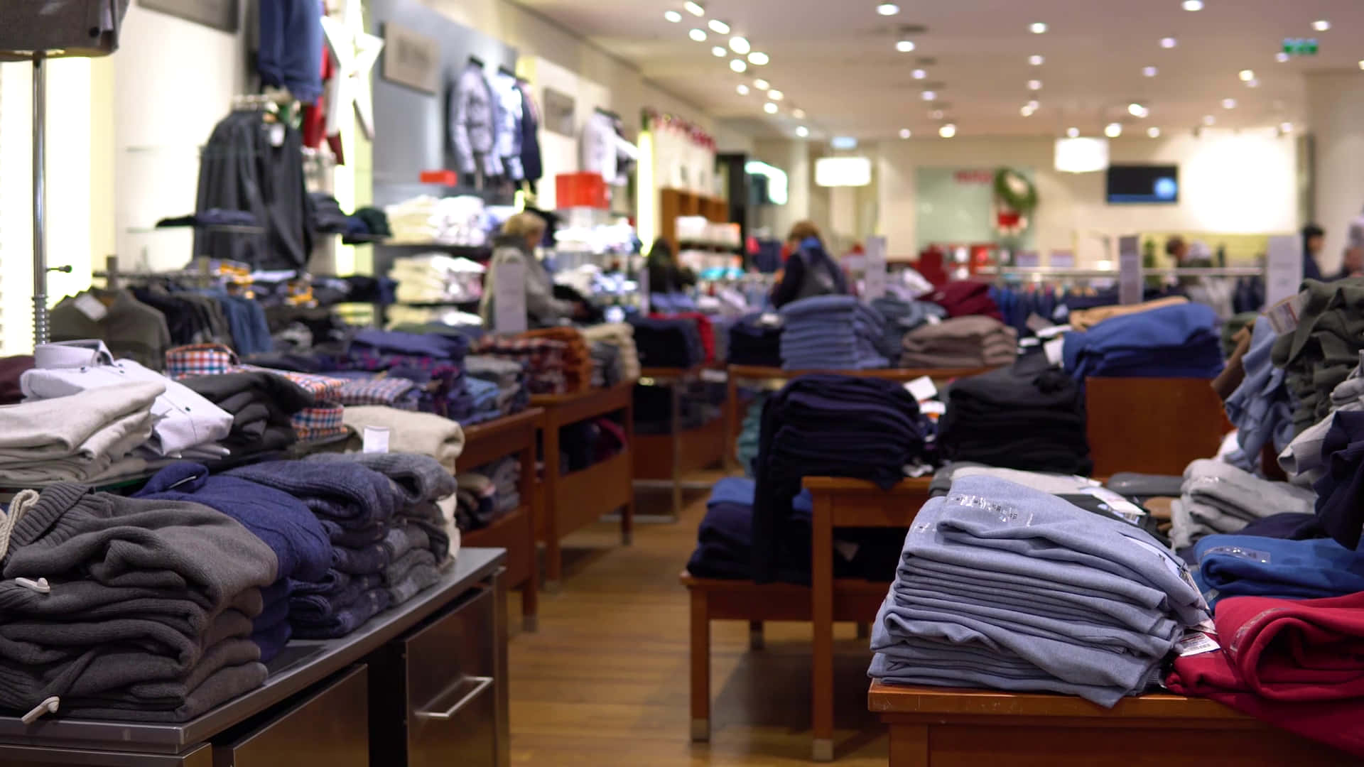 Shopping 桌布 壁紙 — 一排排整齊折疊的柔和色調衣物充滿了光線充足的零售店，店內有木桌和背景中模糊的購物者。