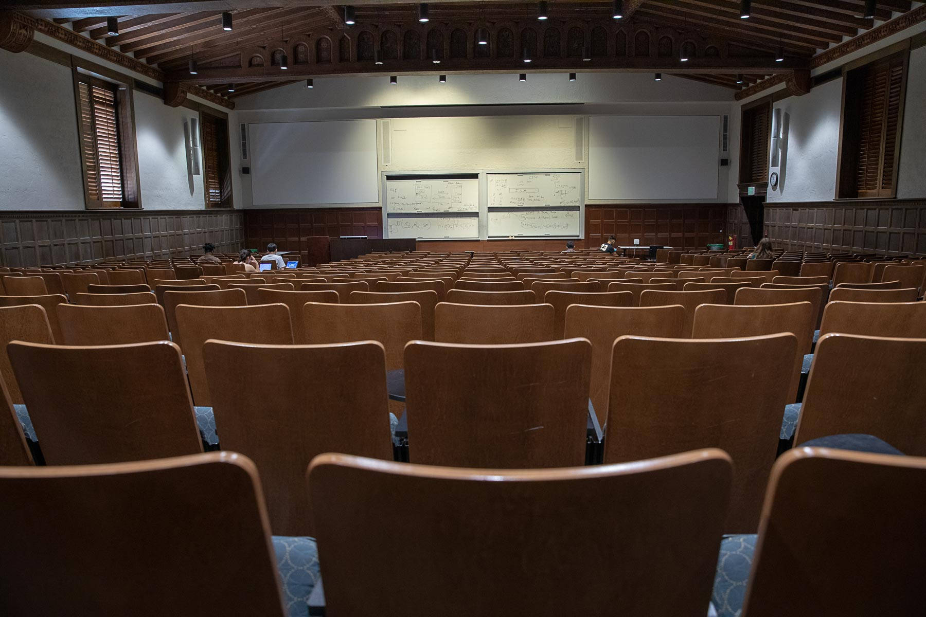 ucla 桌布 壁紙 — 一排排木椅面向設有白板的舞台，位於光線昏暗、有裸露橫樑的演講廳。