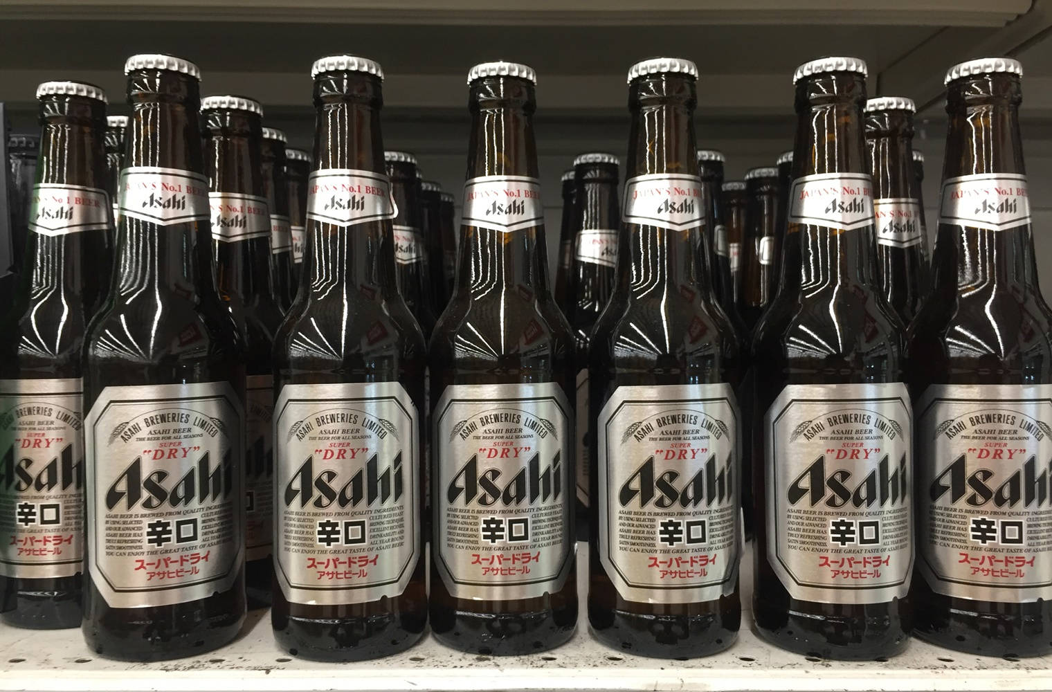 Beer 桌布 壁紙 — 一排排深色玻璃的Asahi Super Dry啤酒瓶整齊地排列在商店貨架上，在明亮的螢光燈下。