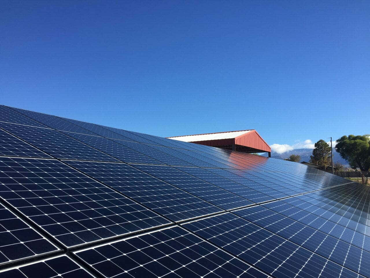 電力 桌布 壁紙 — 一排排深藍色太陽能板在清澈的藍天下閃耀，背景有紅色穀倉和遠山。