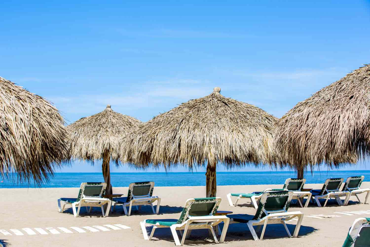 Mexican Beach 桌布 壁紙 — 一排排白色躺椅在草傘下排列在陽光明媚的沙灘上，面向碧綠色的海洋和清澈的藍天。