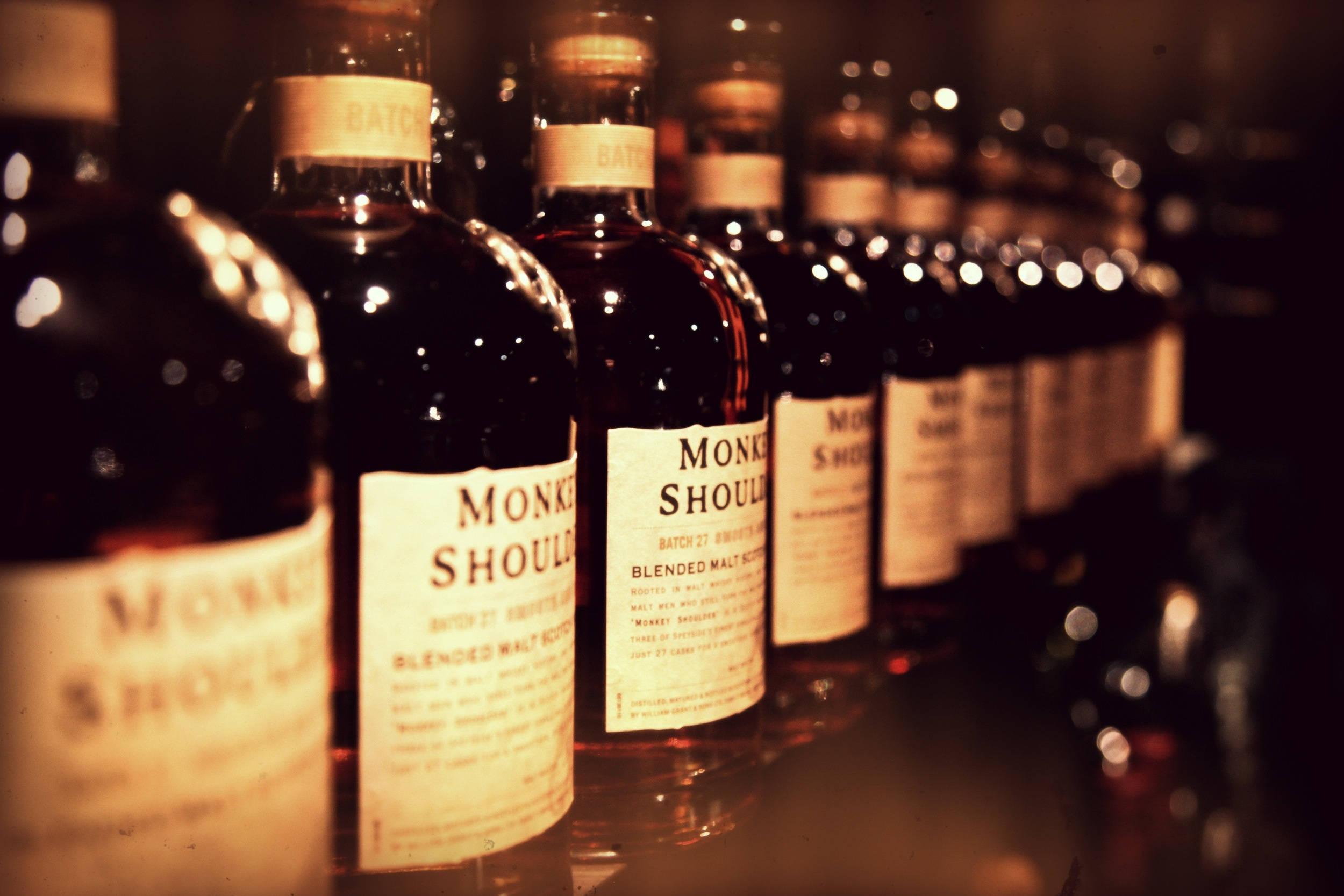 alcohol 桌布 壁紙 — 一排排的Monkey Shoulder威士忌酒瓶在溫暖燈光下閃爍，景深較淺。