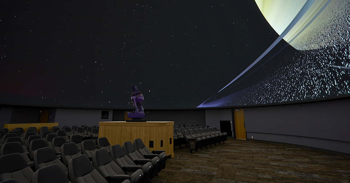 Planetarium 桌布 壁紙 — 一排排空置的劇院座位面向一個彎曲的圓頂，顯示著一個帶有發光行星的星空夜空。