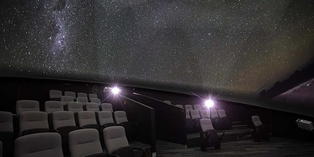 Planetarium 桌布 壁紙 — 一排排空蕩蕩的劇院座位面向一個弧形天花板，顯示出充滿活力的星空和銀河。