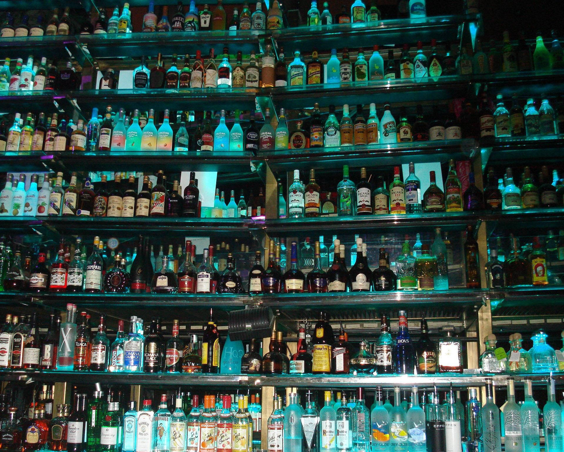 alcohol 桌布 壁紙 — 一排排色彩繽紛的酒瓶在昏暗酒吧的玻璃架上，於霓虹燈下閃耀。