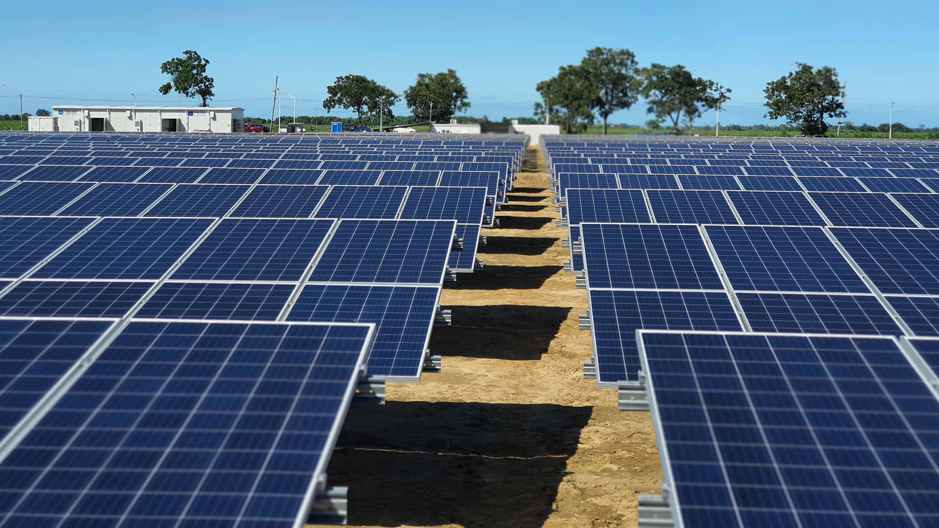 Solar Panel 桌布 壁紙 — 一排排藍色太陽能板在陽光明媚的田野上延伸，天空清澈湛藍，遠處有樹木和一棟小建築。