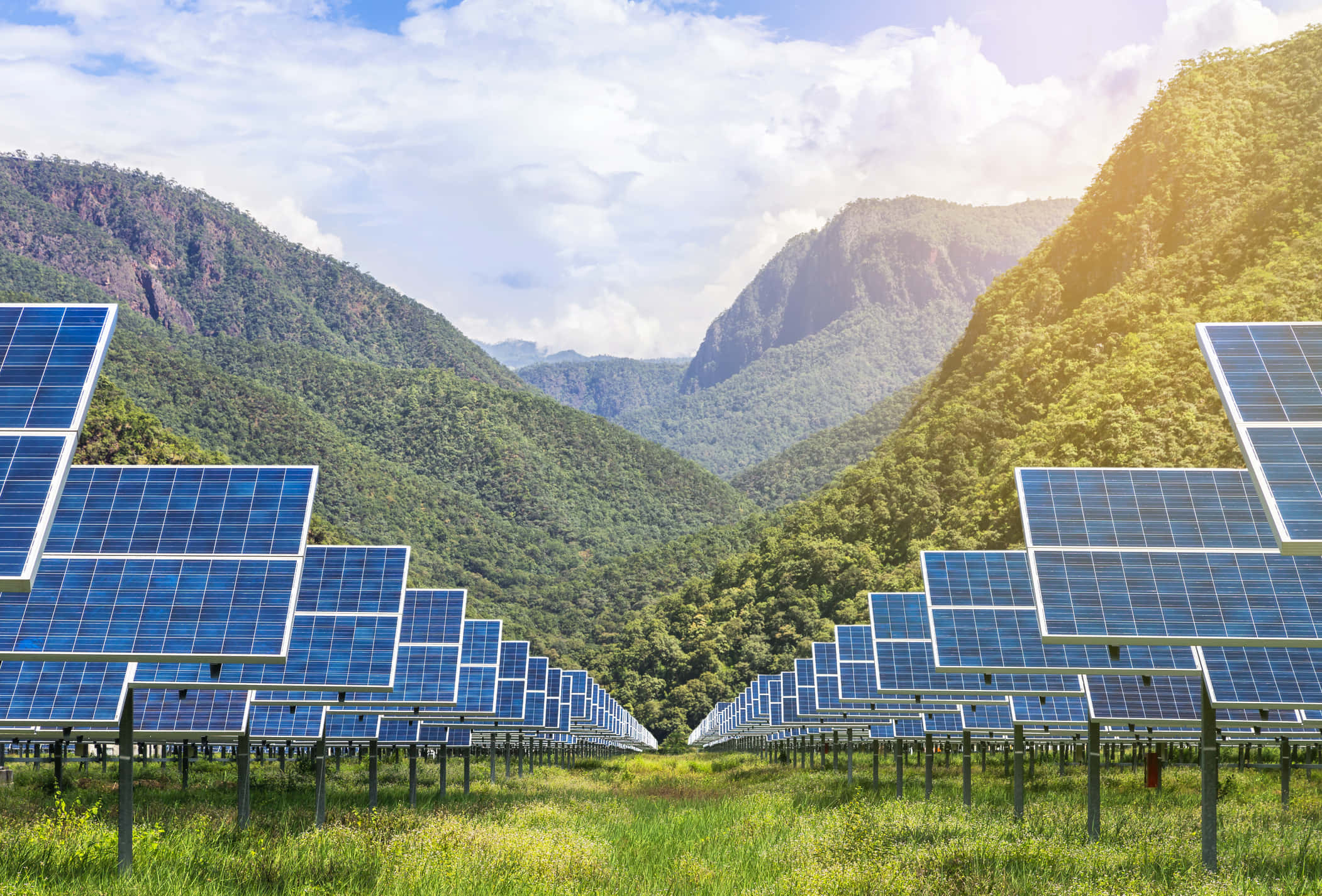 Solar Panel 桌布 壁紙 — 一排排藍色太陽能板延伸穿過一個草地山谷，兩側是茂密的綠色山脈，天空明亮且部分多雲。