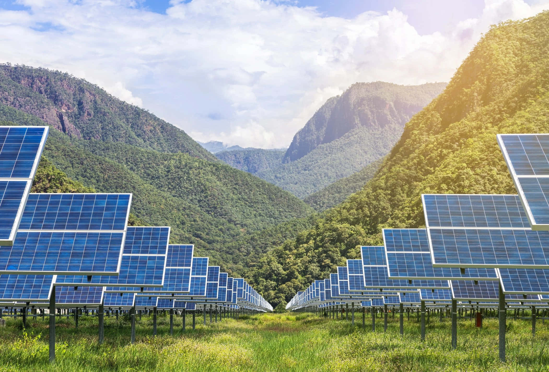 Energy 桌布 壁紙 — 一排排藍色太陽能板延伸穿過一個草地山谷，兩側是茂密的綠色山脈，天空明亮，部分多雲。