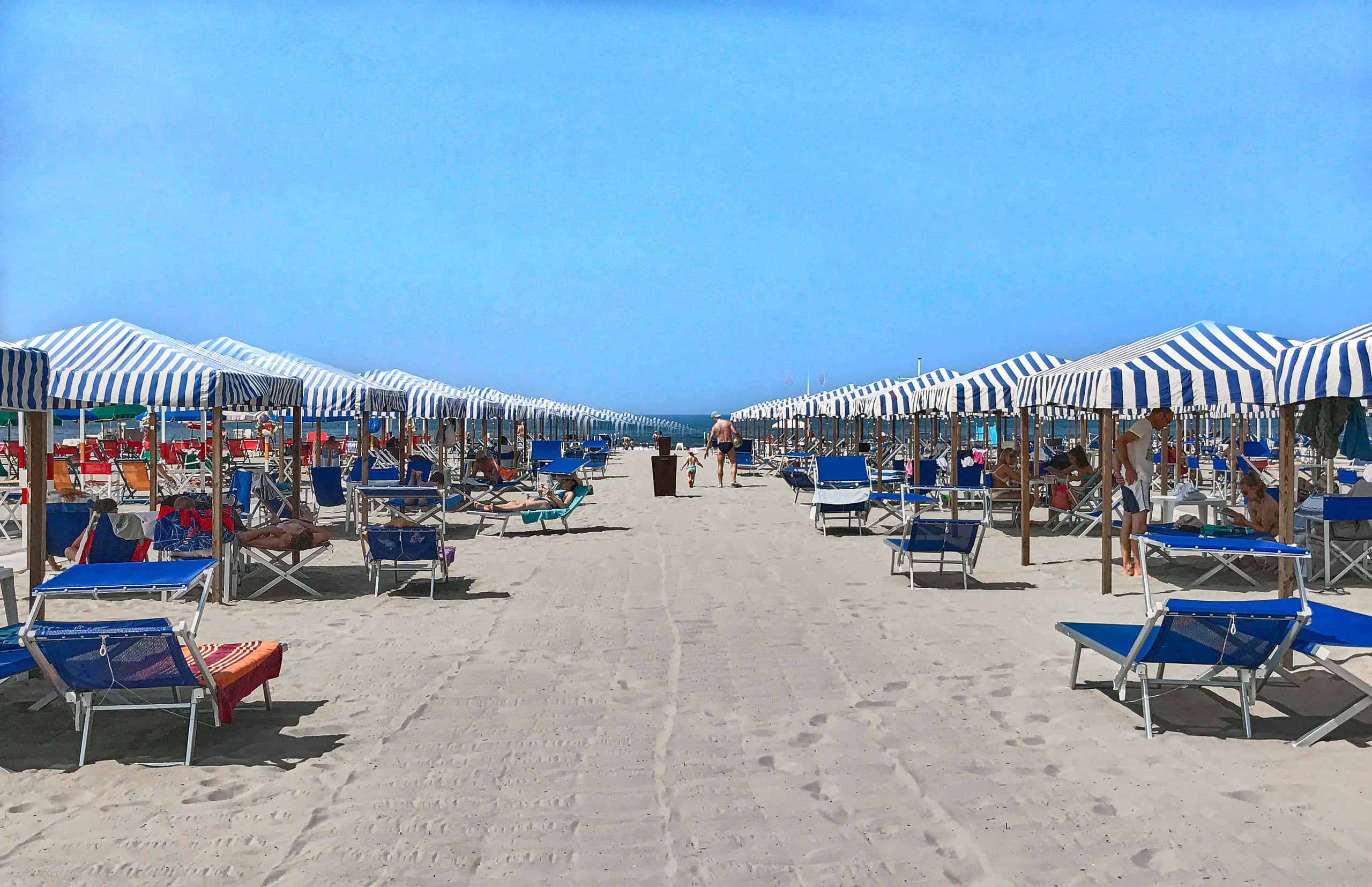 Italian Beach 桌布 壁紙 — 一排排藍色沙灘椅在條紋遮陽傘下，排列在晴朗藍天下的沙灘上。