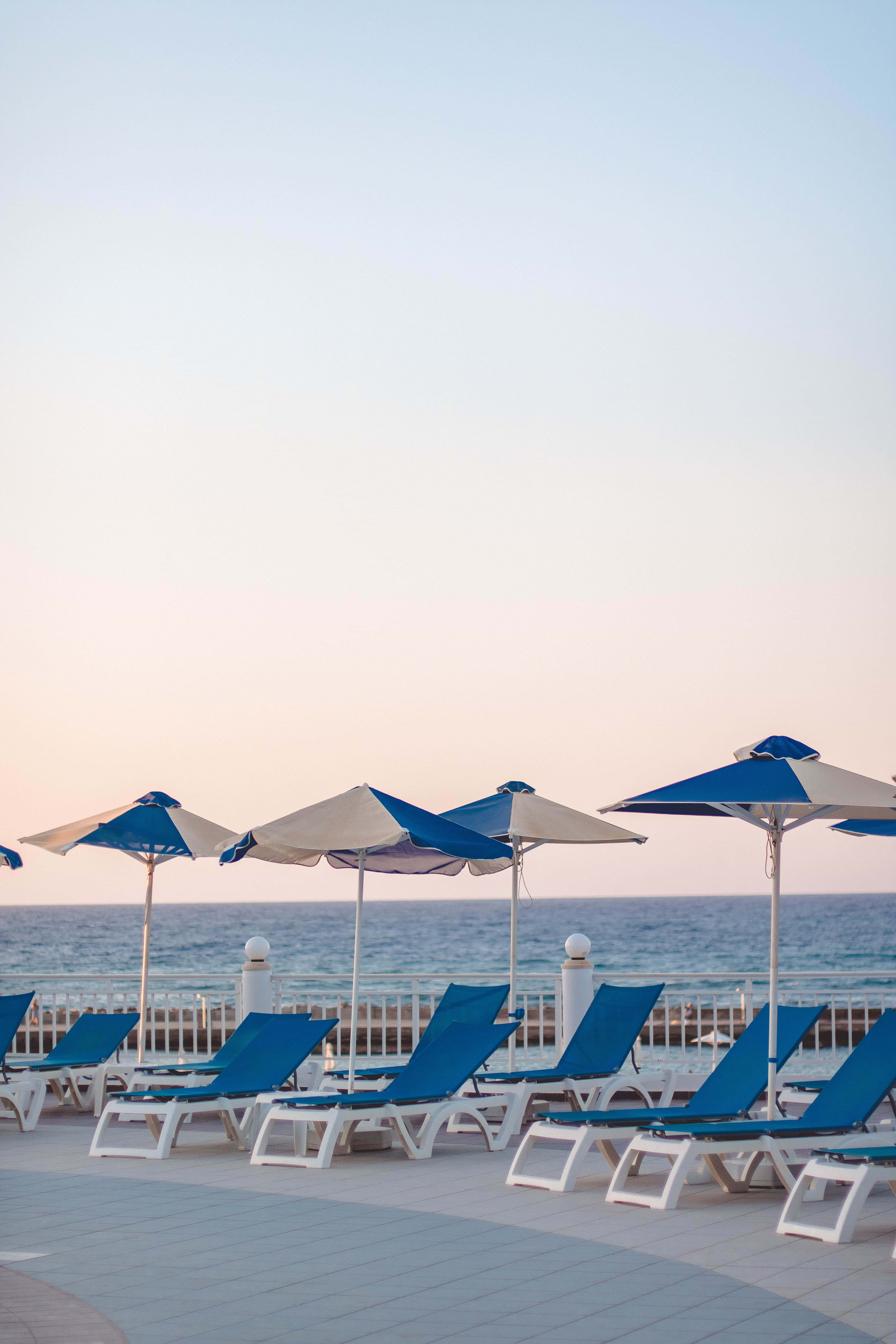Beach Sunbed 桌布 壁紙 — 一排排藍色躺椅和遮陽傘空置在瓷磚甲板上，俯瞰日落時平靜的大海。