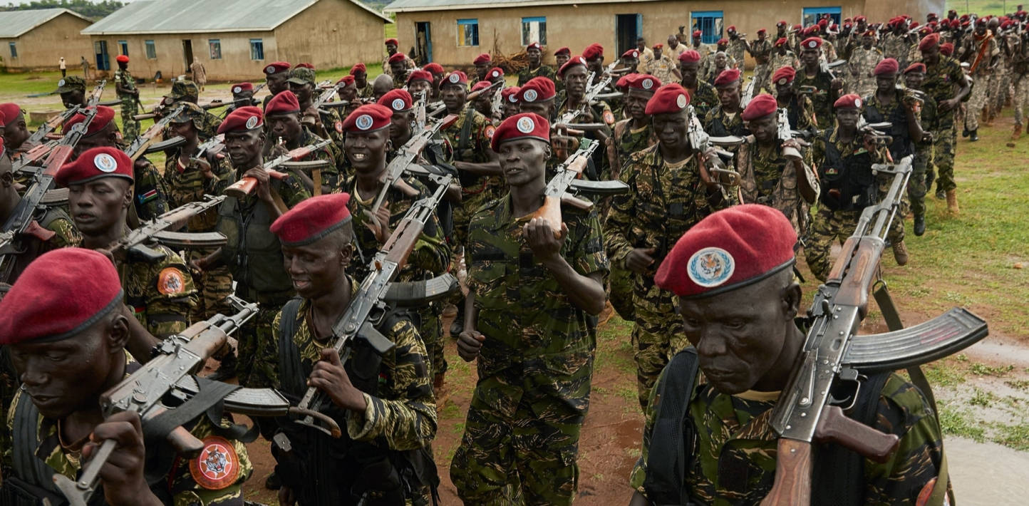 South Sudan 桌布 壁紙 — 一排排身穿迷彩服和紅色貝雷帽的士兵在戶外簡陋建築物附近持步槍列隊行進。