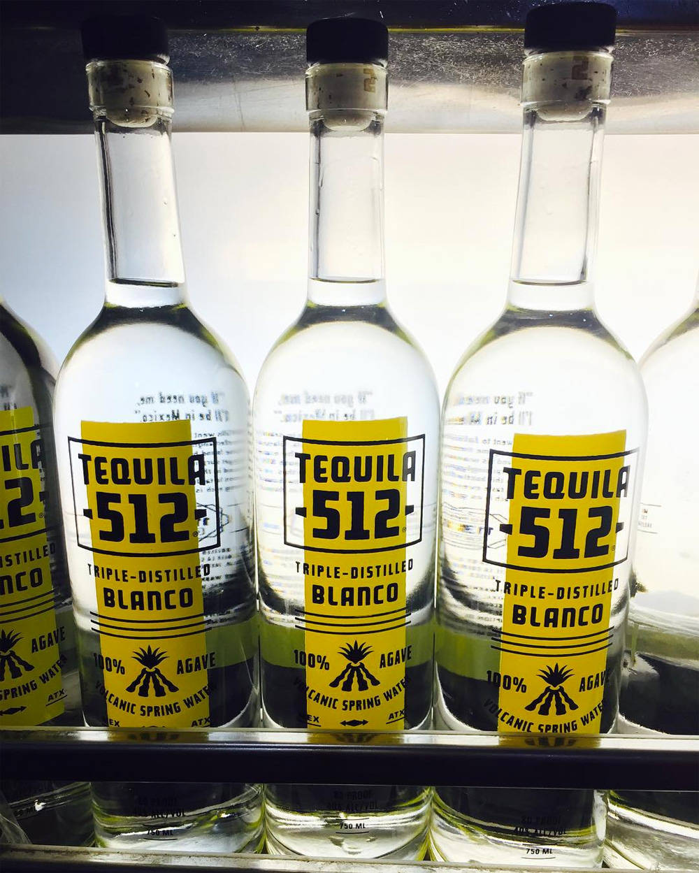 drinks 桌布 壁紙 — 一排排透明龍舌蘭酒瓶，帶有醒目的黃色標籤，整齊地擺放在明亮的燈光下的冷藏架上。