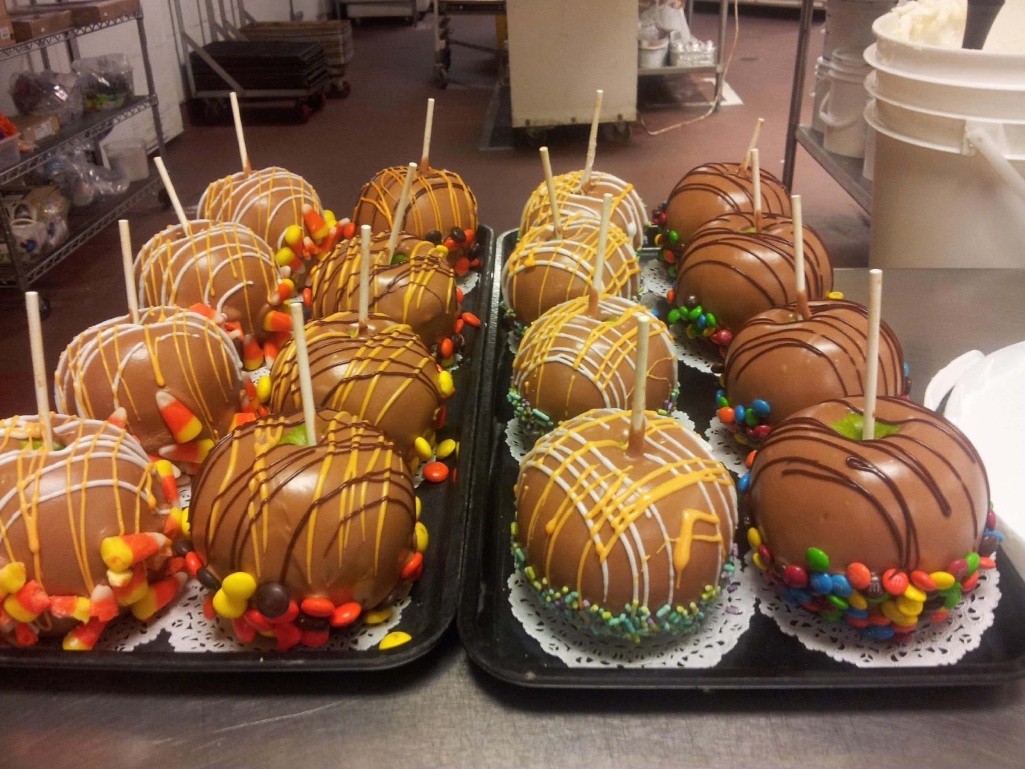 Candy Apples 桌布 壁紙 — 一排排閃亮的焦糖蘋果，裝飾著糖果玉米和色彩繽紛的M&Ms，放在商業廚房的托盤上。
