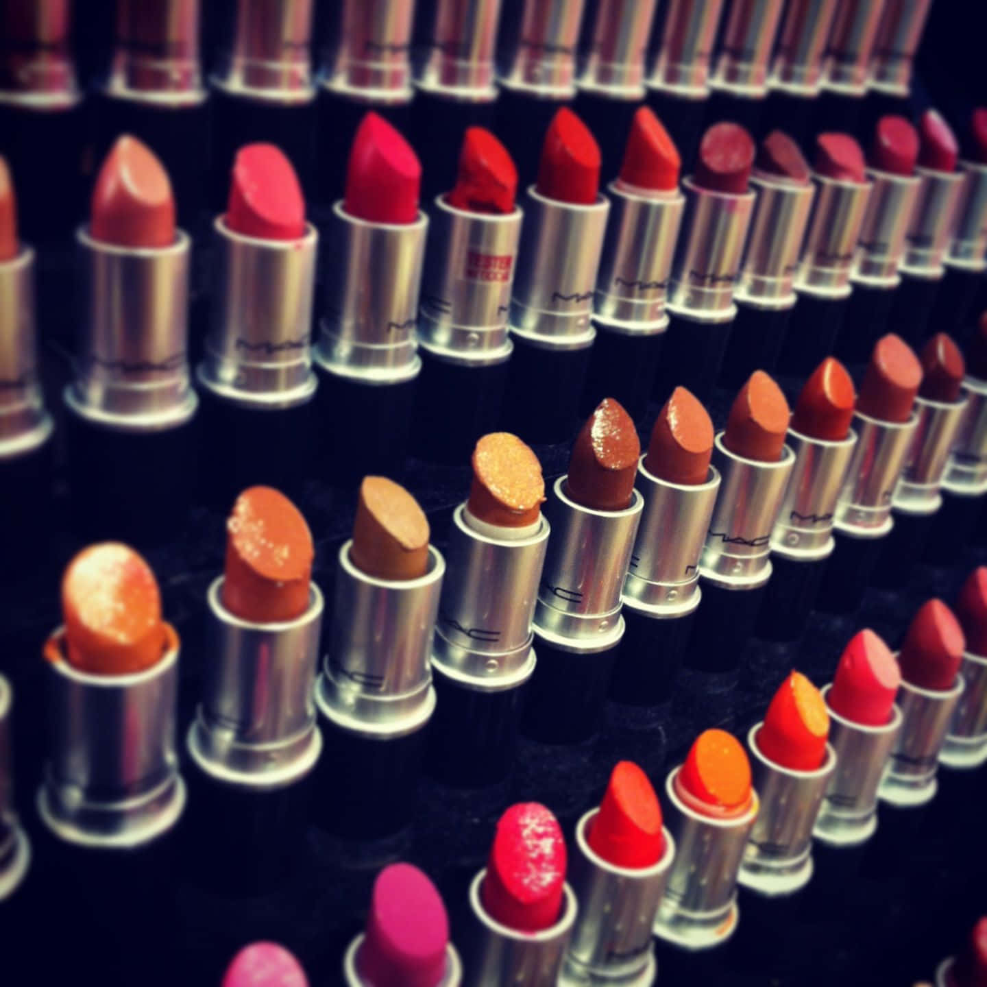Mac Cosmetics 桌布 壁紙 — 一排排鮮紅色、橙色和粉紅色的MAC唇膏在深色表面上對角排列。