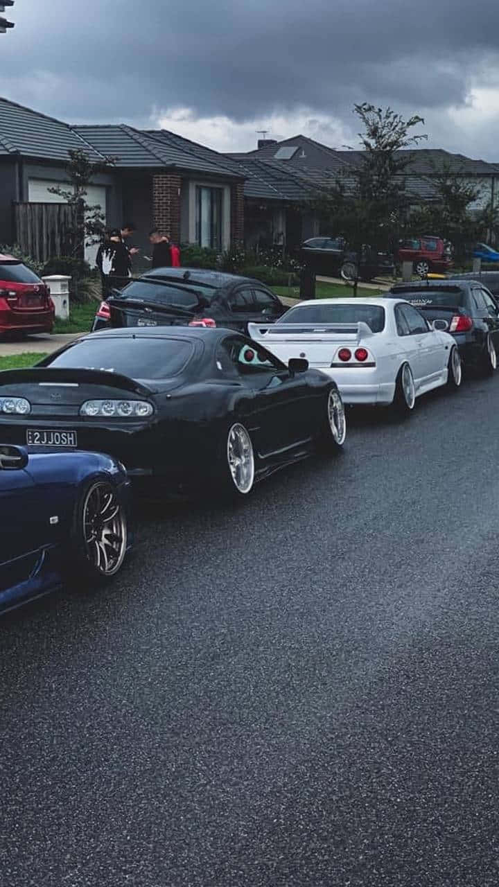 美學 桌布 壁紙 — 一排改裝跑車停放在潮濕的郊區街道上，天空灰濛濛，附近有人站立。