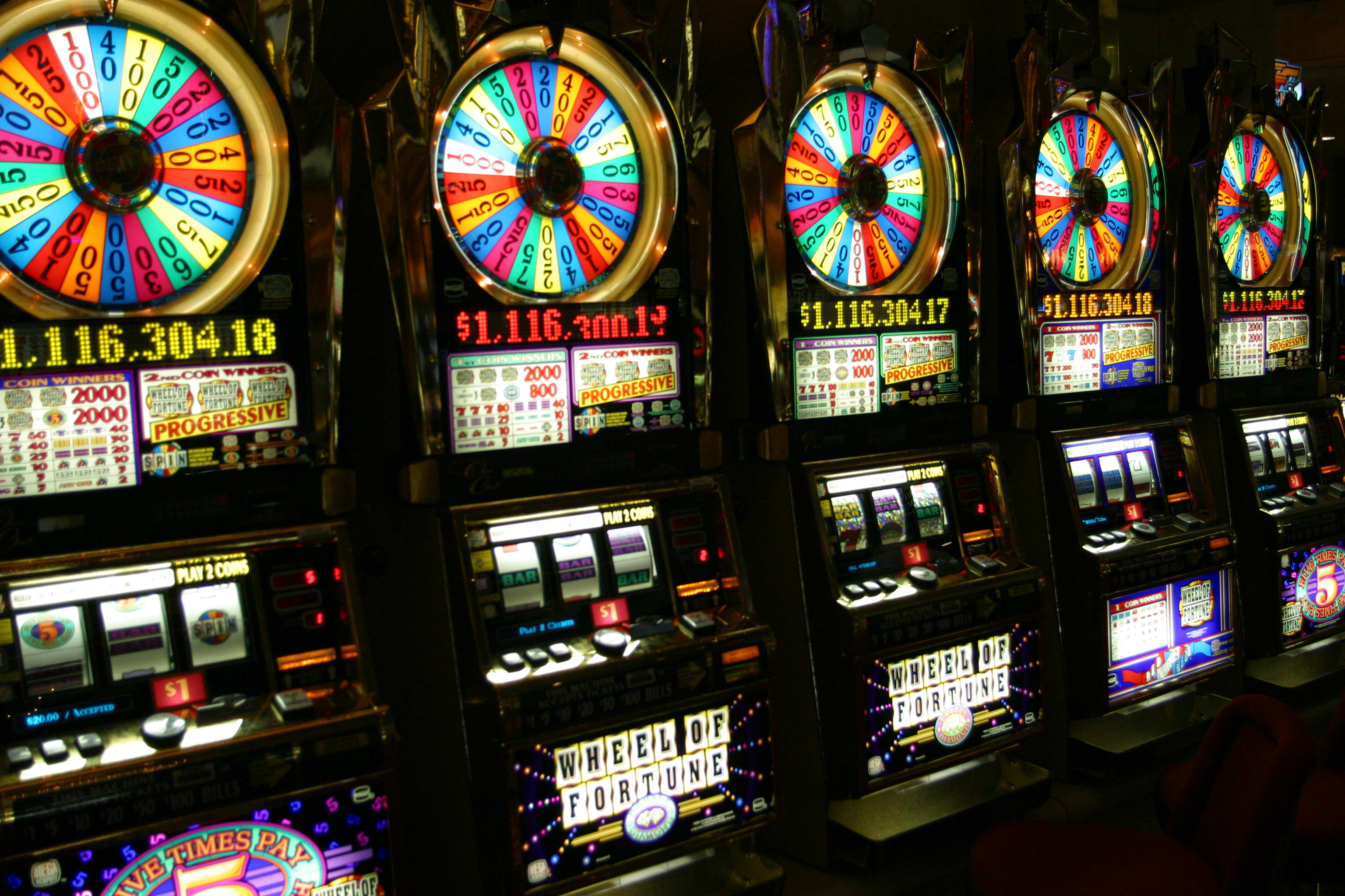 彩色動圖 桌布 壁紙 — 一排明亮照明的 Wheel of Fortune 老虎機在賭場中發光，配有色彩繽紛的旋轉輪盤和累積獎金顯示屏。