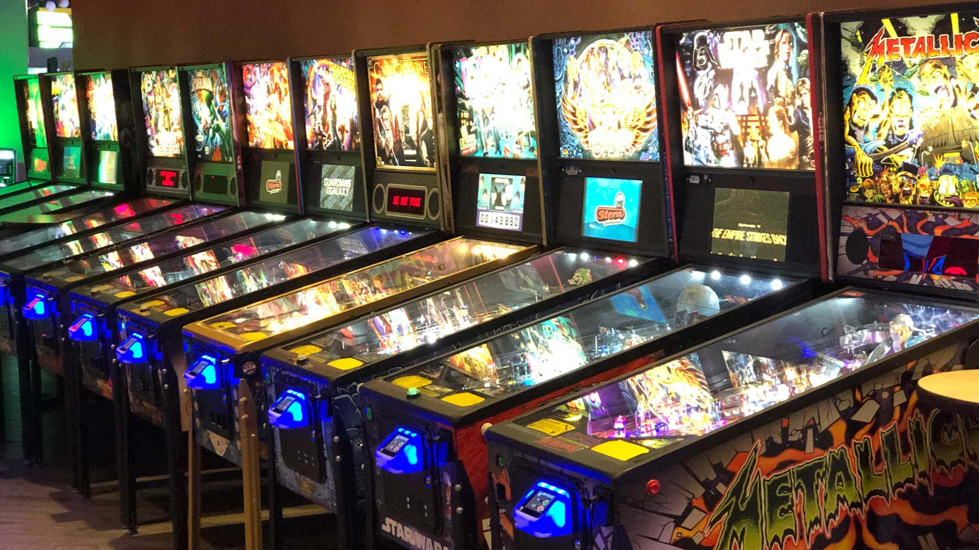 Arcade Games 桌布 壁紙 — 一排明亮照明的復古彈珠台，帶有彩色藝術品和發光顯示屏，位於復古遊戲機中心。