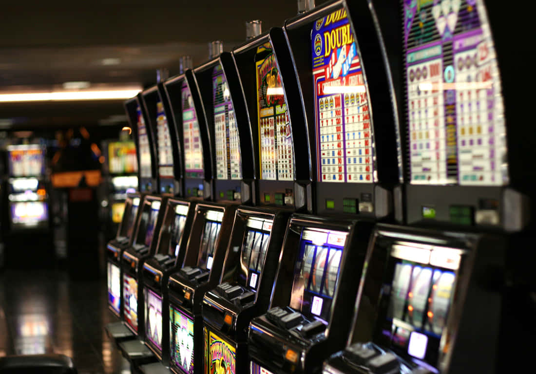 game 桌布 壁紙 — 一排明亮照明的老虎機，位於燈光昏暗的賭場中，帶有發光的螢幕和色彩繽紛的顯示器。