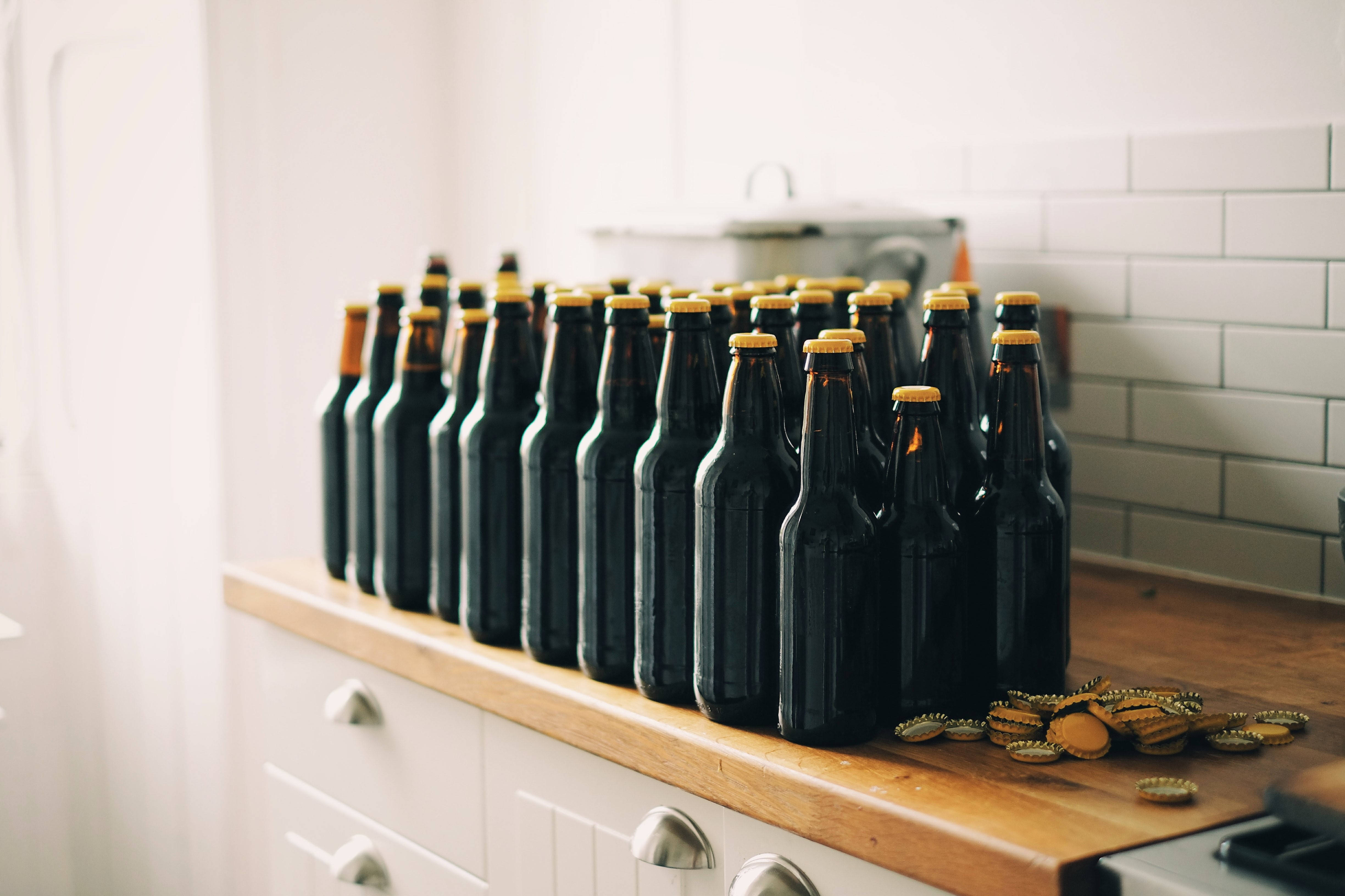 alcohol 桌布 壁紙 — 一排深色啤酒瓶帶金色瓶蓋排列在木製廚房檯面上，旁邊散落著瓶蓋。