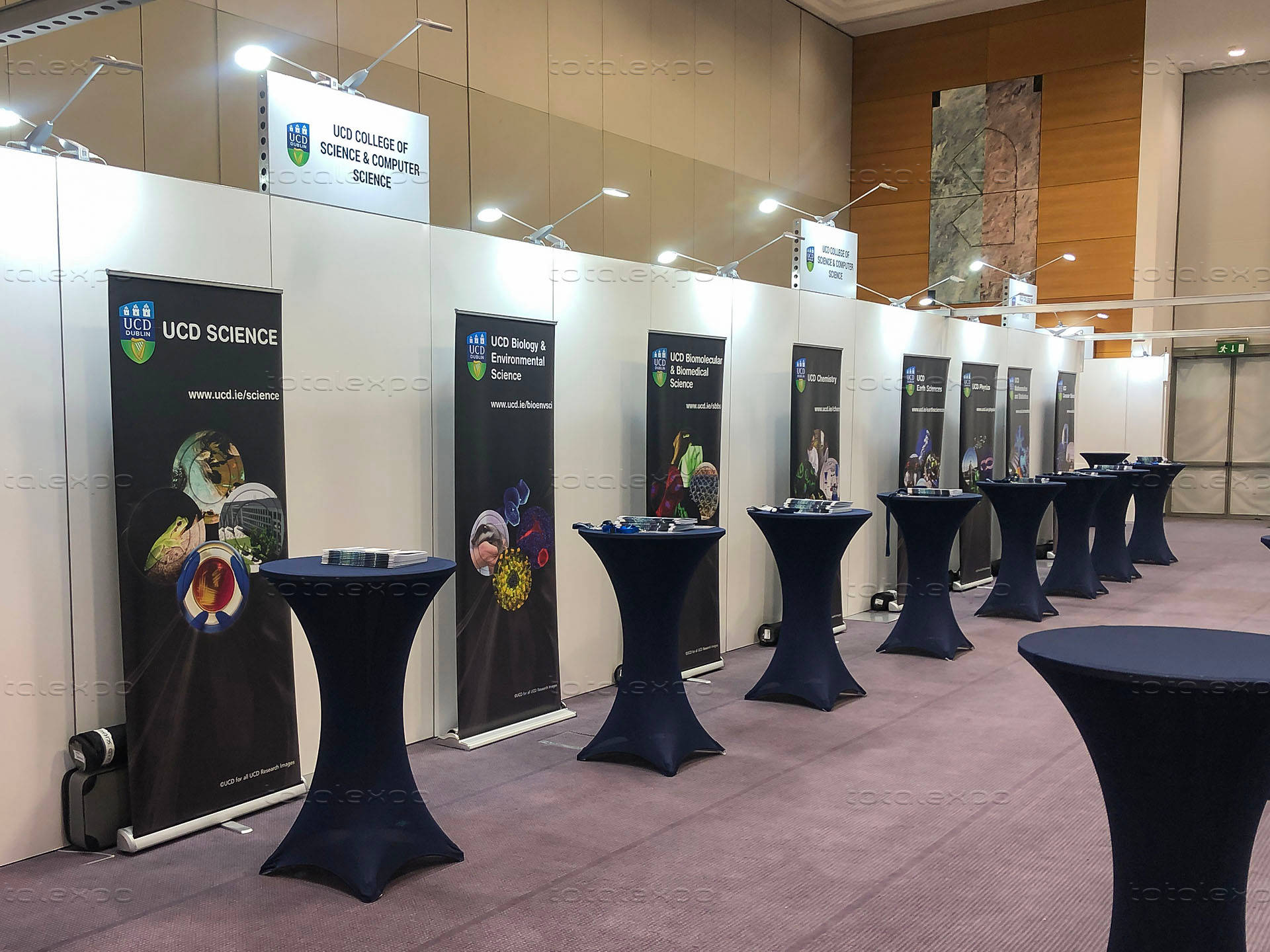 桌布 壁紙 — 一排深藍色高腳桌排列在鋪有地毯的展覽廳中，掛有UCD Science橫幅及天花燈。