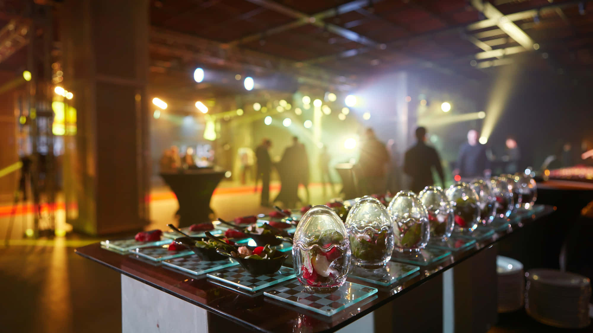 Corporate Event 桌布 壁紙 — 一排玻璃花瓶盛載紅玫瑰，放在格子桌布上，處於燈光昏暗的活動大廳，背景有模糊的賓客和舞台燈光。