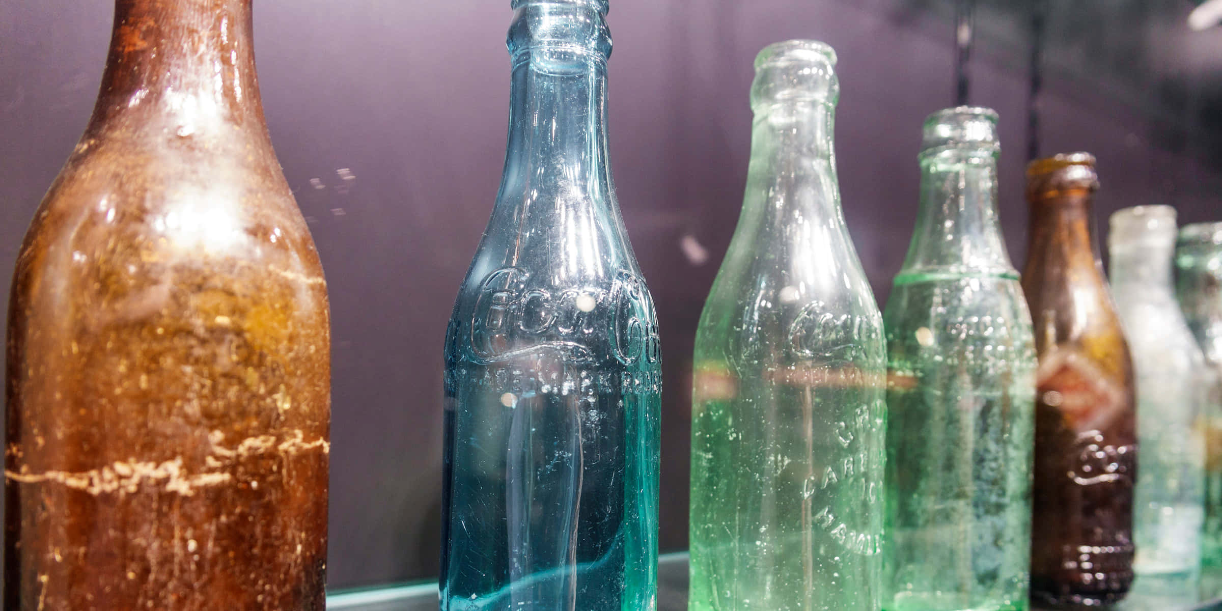 Old Bottles 桌布 壁紙 — 一排琥珀色、藍色和綠色色調的復古玻璃瓶，排列在深紫色背景前。