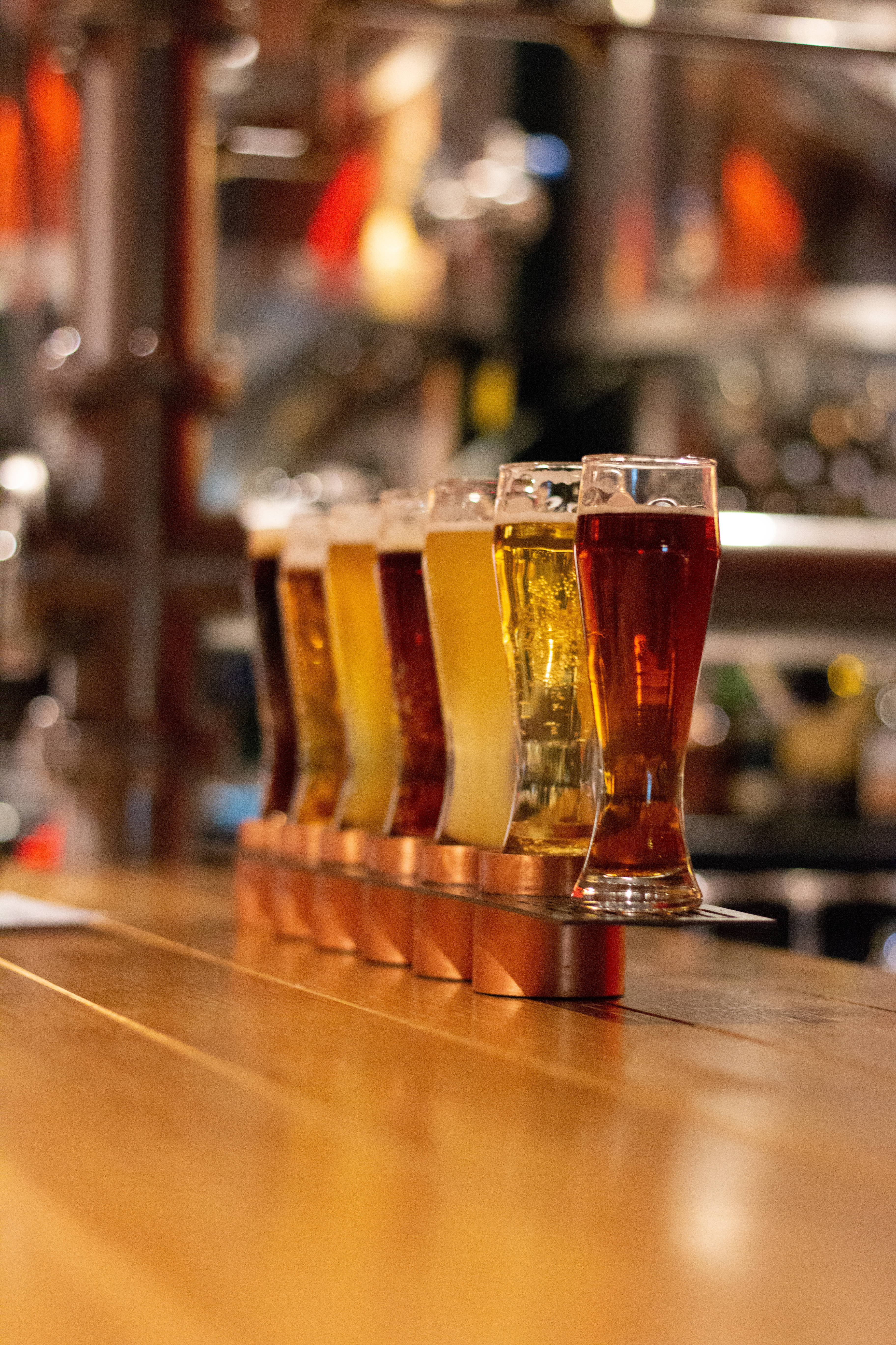 Beer 桌布 壁紙 — 一排琥珀色和金色的啤酒杯整齊地放在拋光木製吧檯上，配有溫暖的環境燈光。