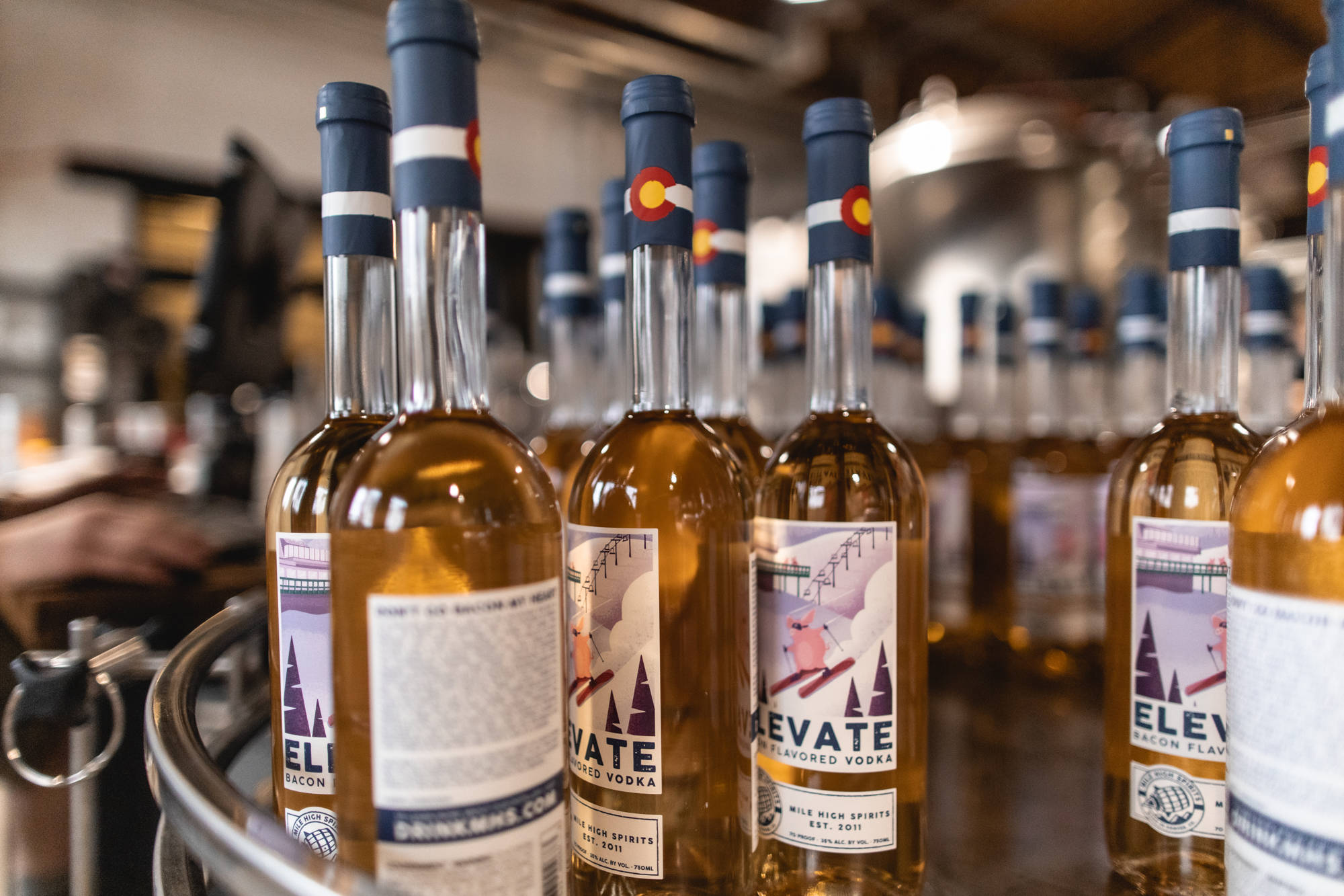 alcohol 桌布 壁紙 — 一排琥珀色培根味伏特加酒瓶，帶有科羅拉多州旗瓶蓋，放在鄉村酒吧環境中的冷凍金屬桶中。