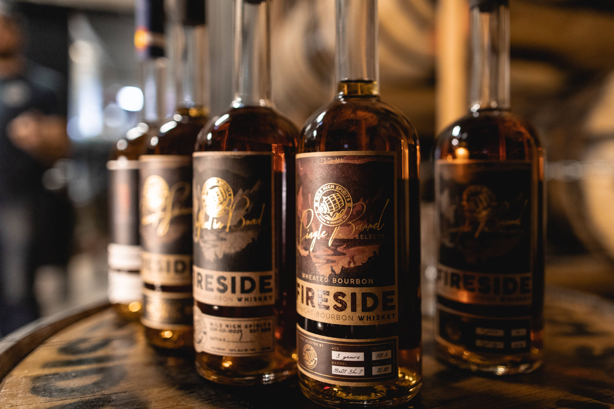 drinks 桌布 壁紙 — 一排琥珀色威士忌酒瓶帶金色標籤，放置在溫暖燈光下的釀酒廠木桶上。