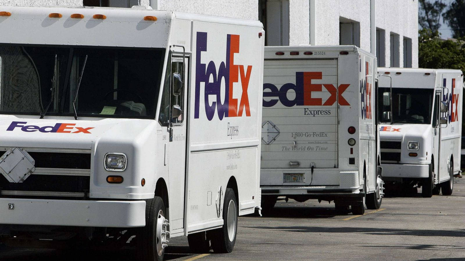 聯邦快遞 桌布 壁紙 — 一排白色FedEx送貨車停在陽光明媚的停車場，背景是晴朗的藍天和建築物。