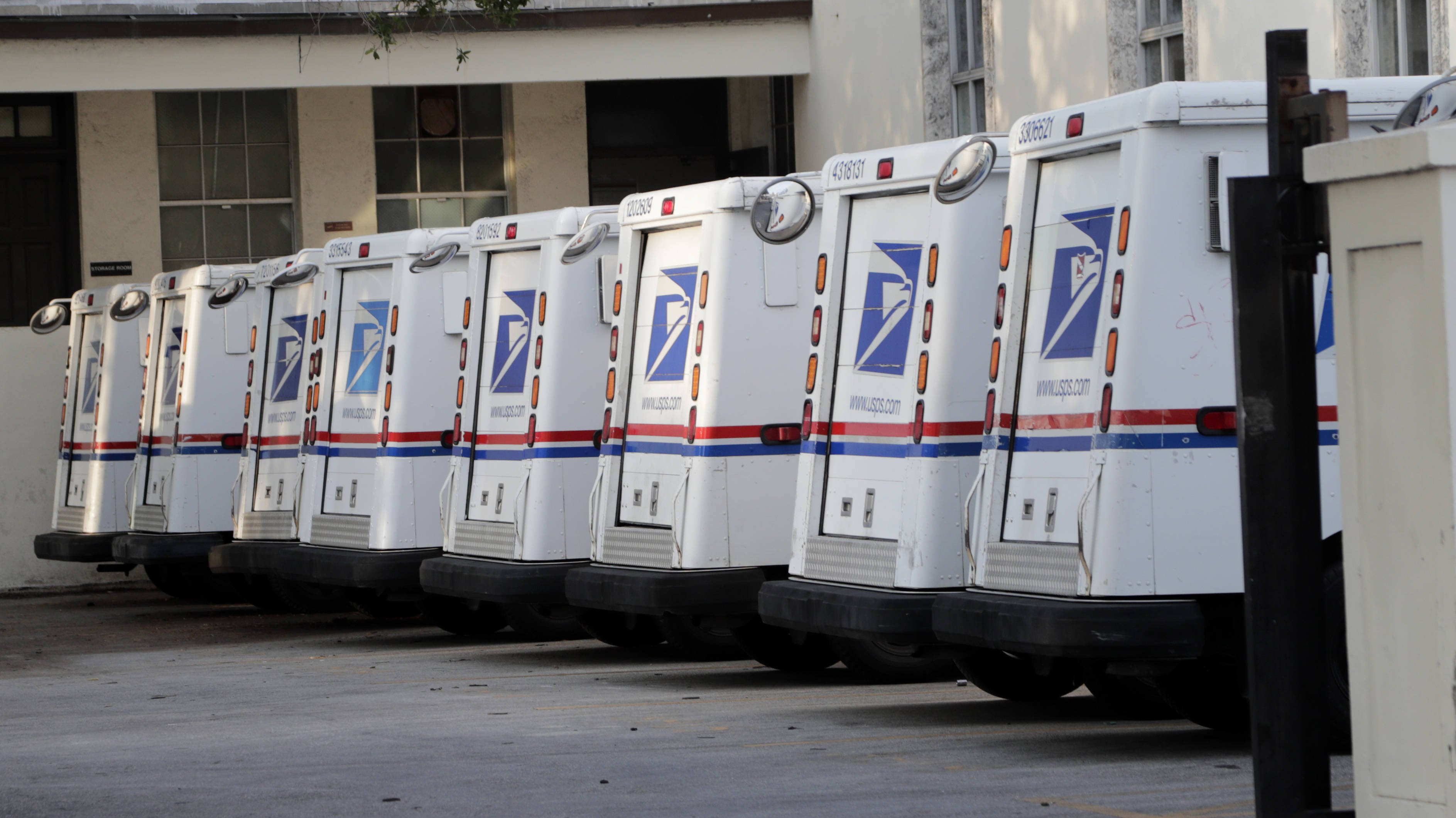 Trucks 桌布 壁紙 — 一排白色USPS郵政貨車停在一棟有明顯磨損的建築物外面。