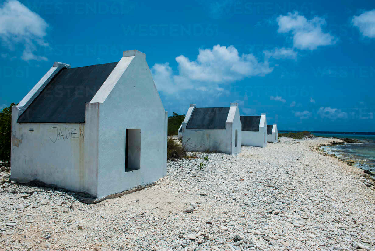 Hut 桌布 壁紙 — 一排白色鮮明的海邊小屋，黑色屋頂，沿著岩石海岸排列，天空湛藍，雲朵蓬鬆。