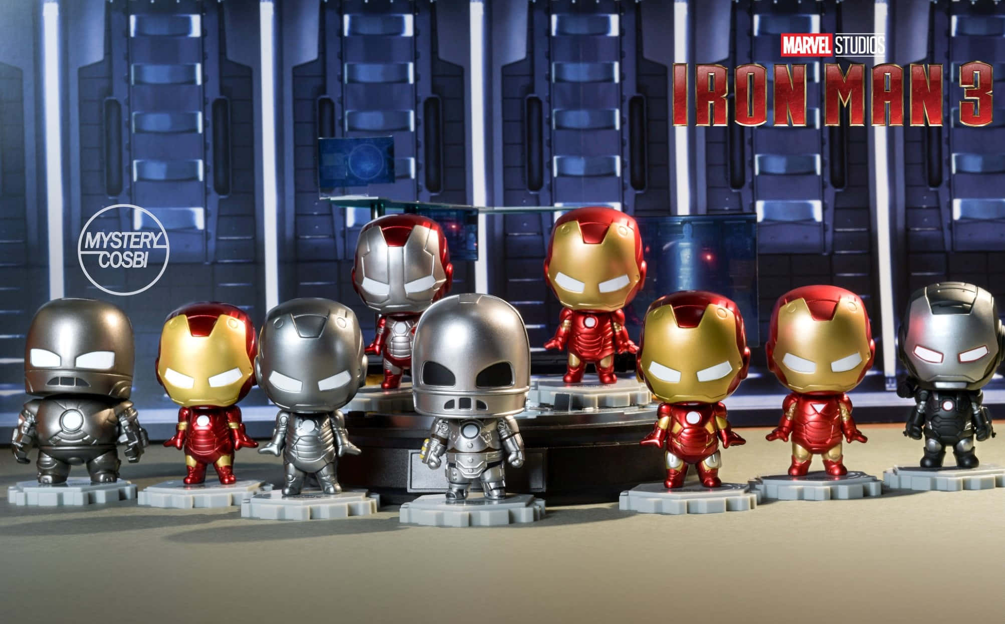 Iron Man Bobbleheads 桌布 壁紙 — 一排穿著不同裝甲服的Q版鋼鐵人公仔，在帶有『鋼鐵人3』品牌標誌的科技主題舞台上展示。