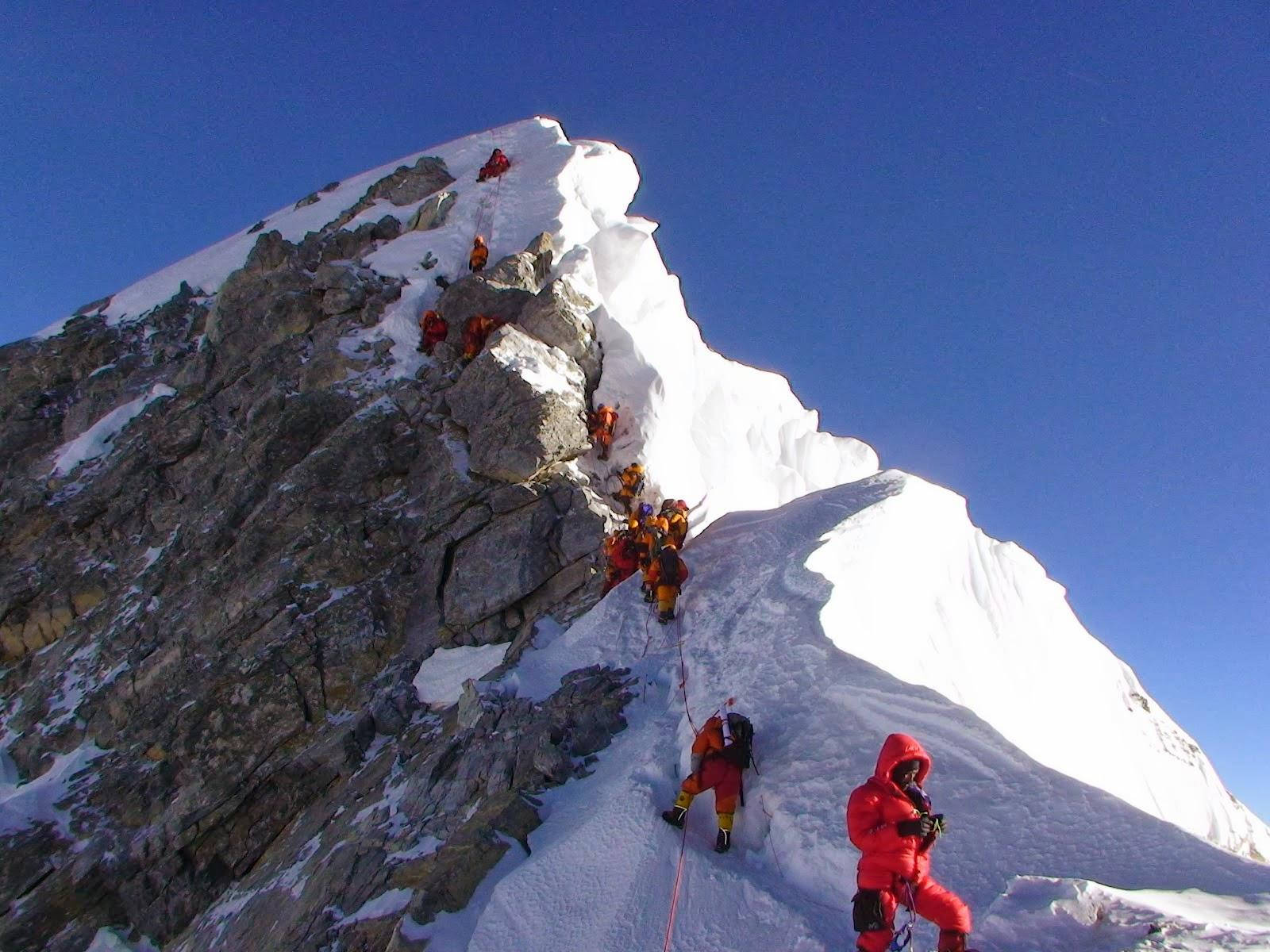 Climbing 桌布 壁紙 — 一排穿著亮橙色套裝的登山者在晴朗的藍天下攀登陡峭的雪岩山脊。
