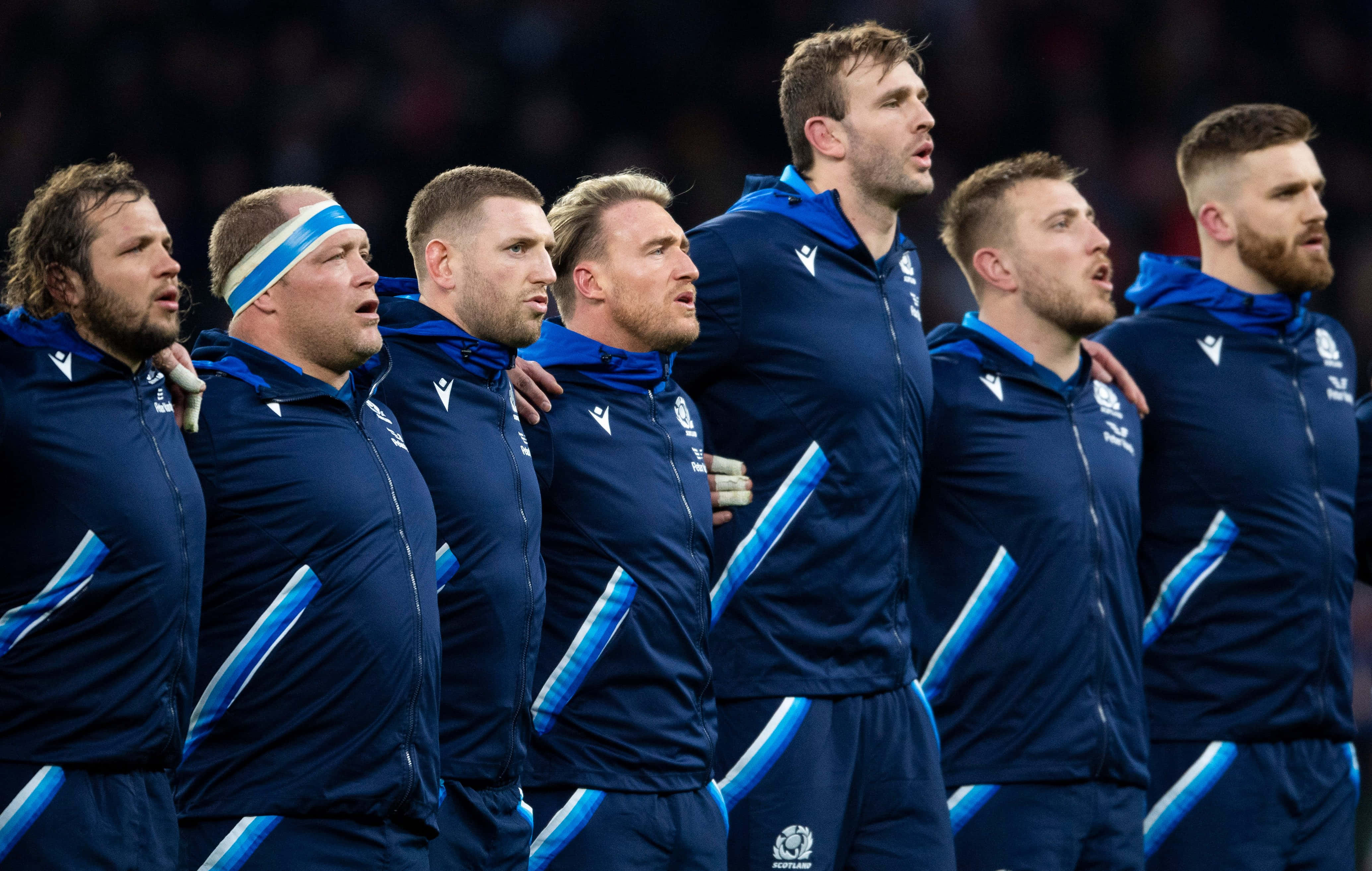 Scotland Rugby 桌布 壁紙 — 一排穿著藍色運動服的橄欖球運動員在國歌演奏期間莊嚴地站著，手臂互相搭在對方肩上。