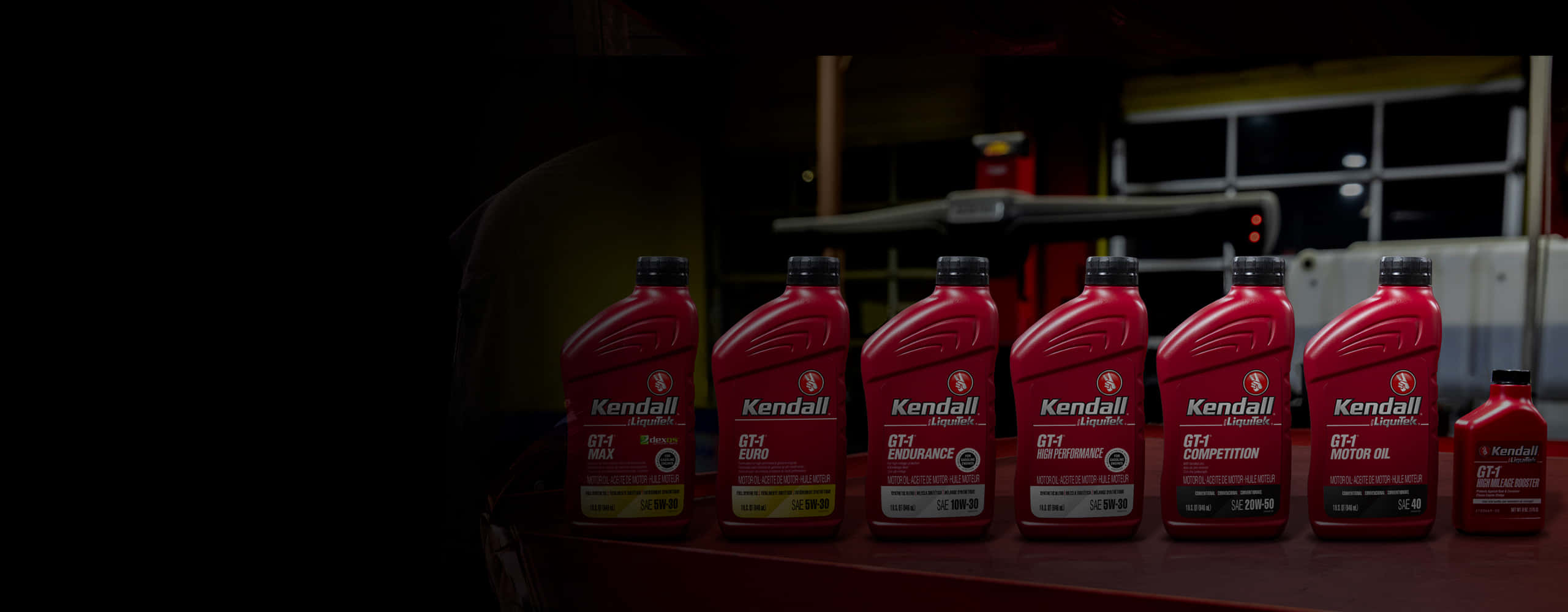 Engine Oil 桌布 壁紙 — 一排紅色Kendall機油瓶整齊排列在昏暗車庫的架子上，背景中有一位技工。