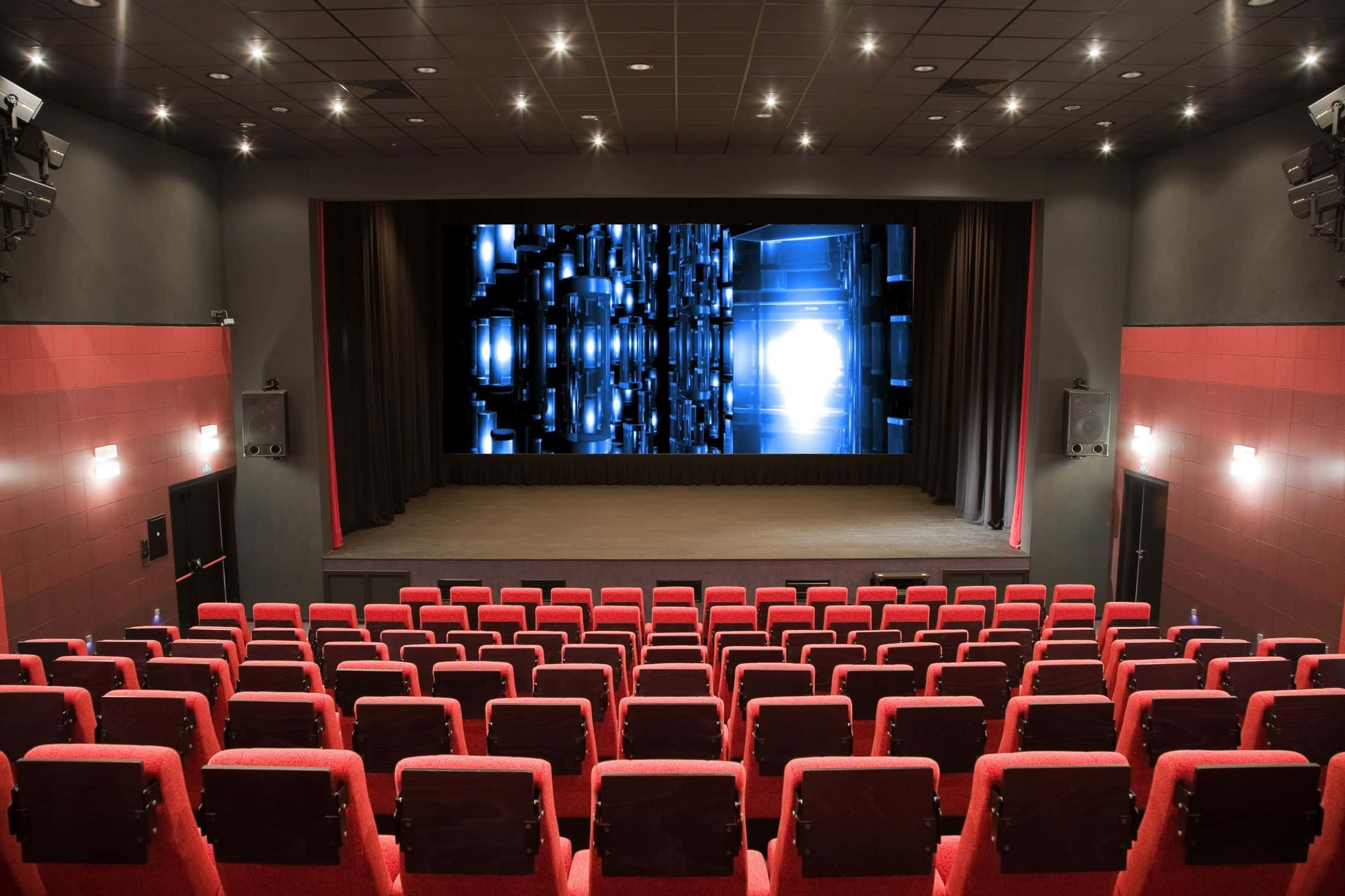 Theater 桌布 壁紙 — 一排紅色電影院座位面向現代劇院中發光的藍色數位螢幕，天花板設有嵌入式燈光。