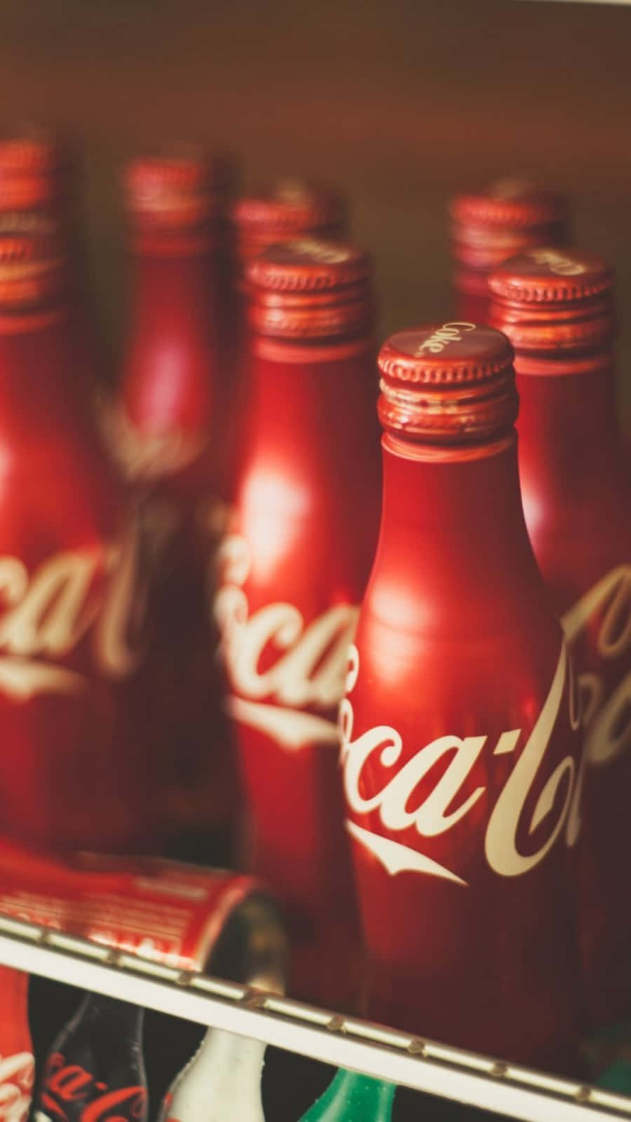 coca cola 桌布 壁紙 — 一排經典的紅色可口可樂玻璃瓶，帶有白色字樣，整齊地放在冰箱架上，柔和地以暖色調照明。