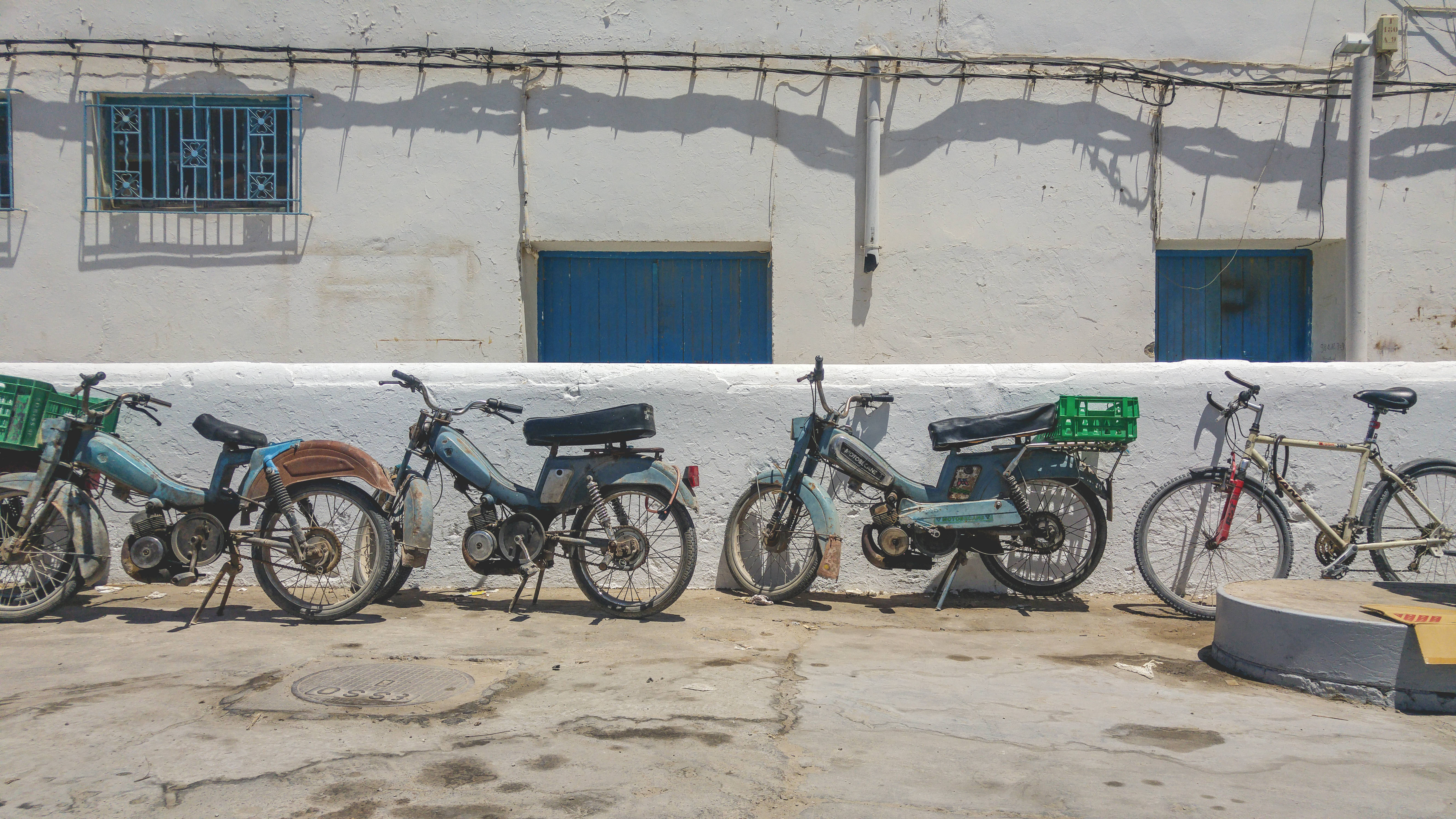 Tunisia 桌布 壁紙 — 一排經風吹日曬的機車和一輛腳踏車，停靠在陽光照射的白色牆壁旁，牆上有藍色窗戶。