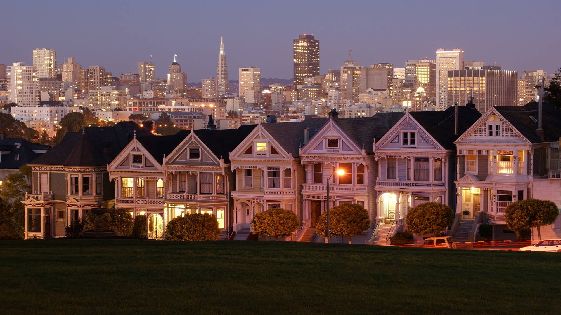 San Francisco HD 桌布 壁紙 — 一排維多利亞式房屋在黃昏時溫暖地發光，背後是舊金山燈火通明的天際線。