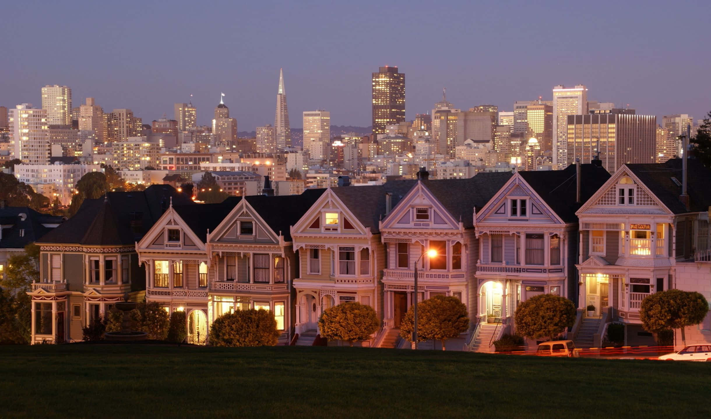 2440x1440 San Francisco 桌布 壁紙 — 一排維多利亞式房屋在黃昏時溫暖地亮起，背後是舊金山閃爍的天際線。